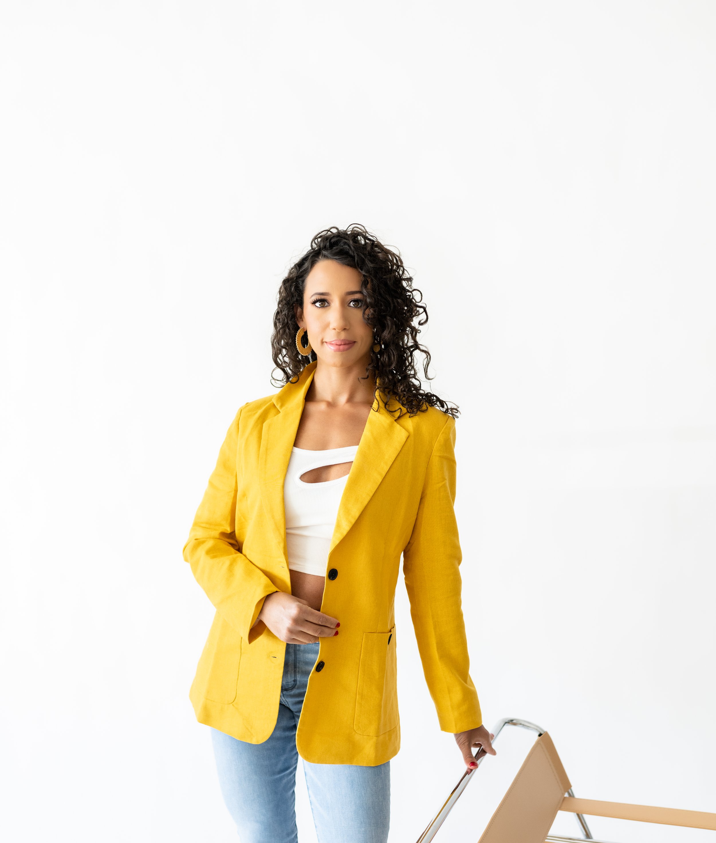 Woman with curly dark hair wearing a yellow blazer, white cutout top, and light blue jeans standing next to a beige luggage trolley against a white background.