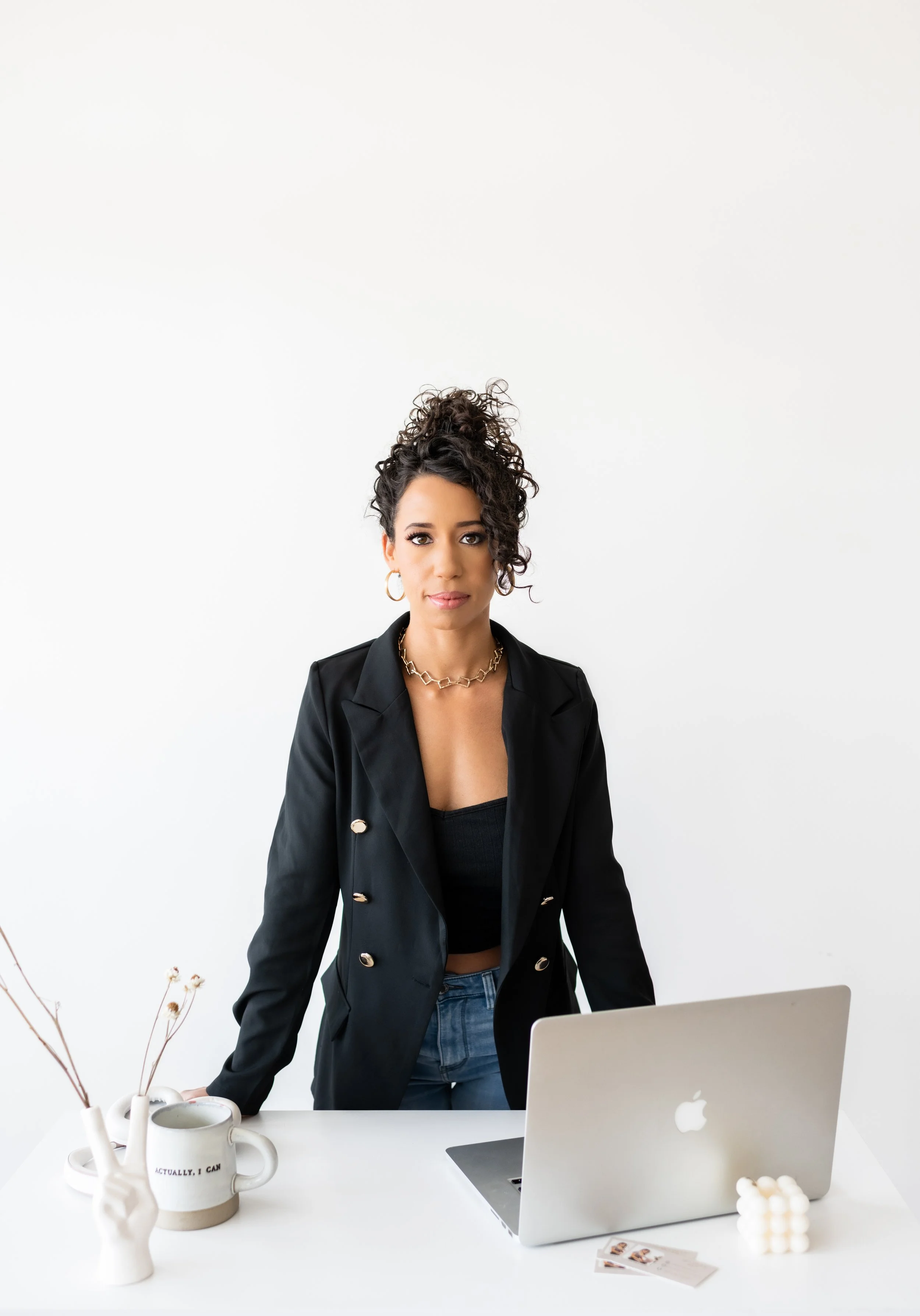 Woman with curly hair standing behind a white desk, wearing a black blazer, black top, and gold jewelry, in front of a silver laptop, with a white mug, a vase with dried branches, and some cards on the desk.