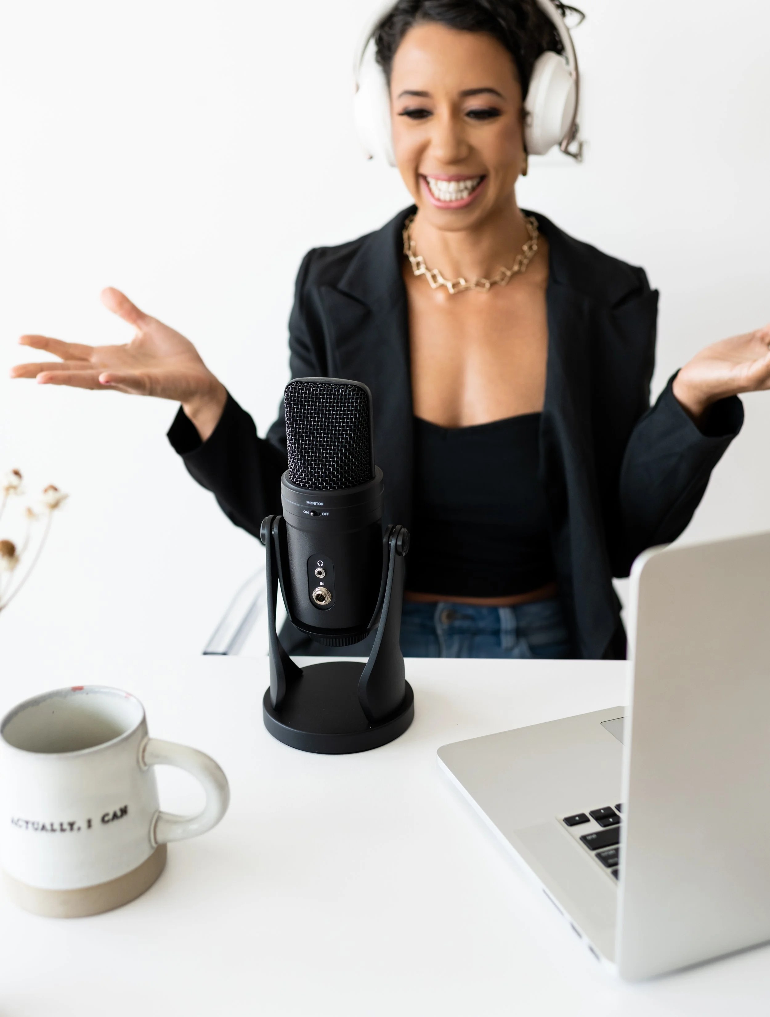 A woman with headphones speaking into a microphone during a podcast recording session, with a laptop and a mug on the table.