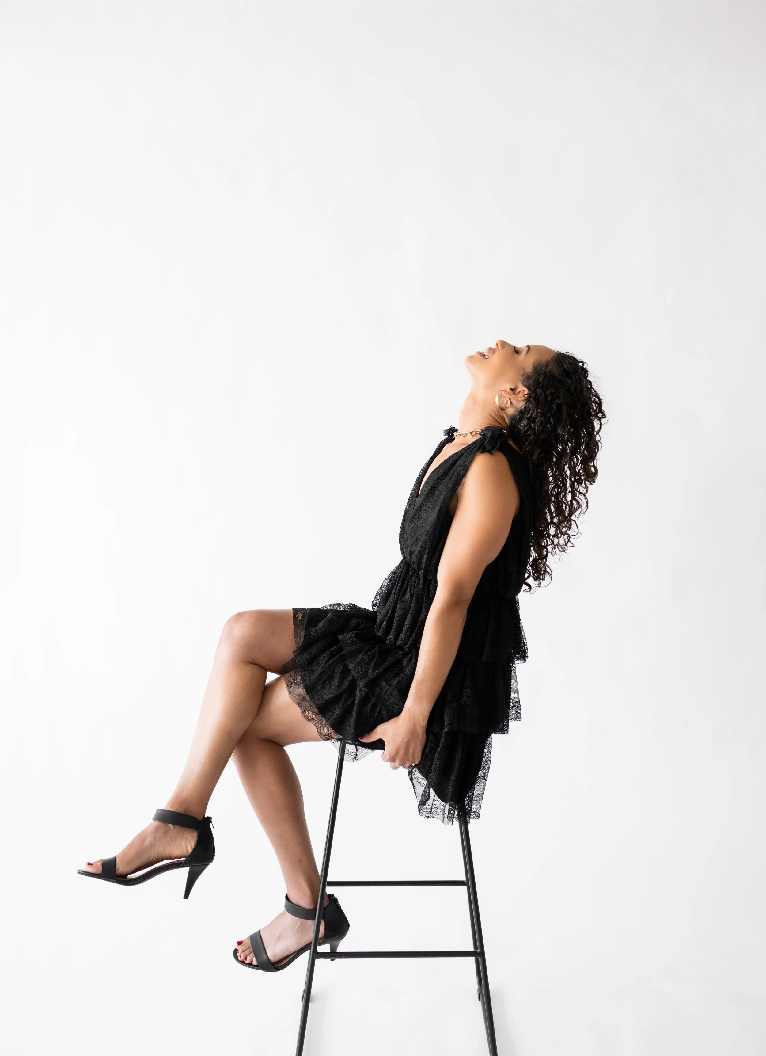 Woman sitting on a black stool with legs crossed, wearing a black lace dress and high heels, smiling with head tilted back against a plain white background.