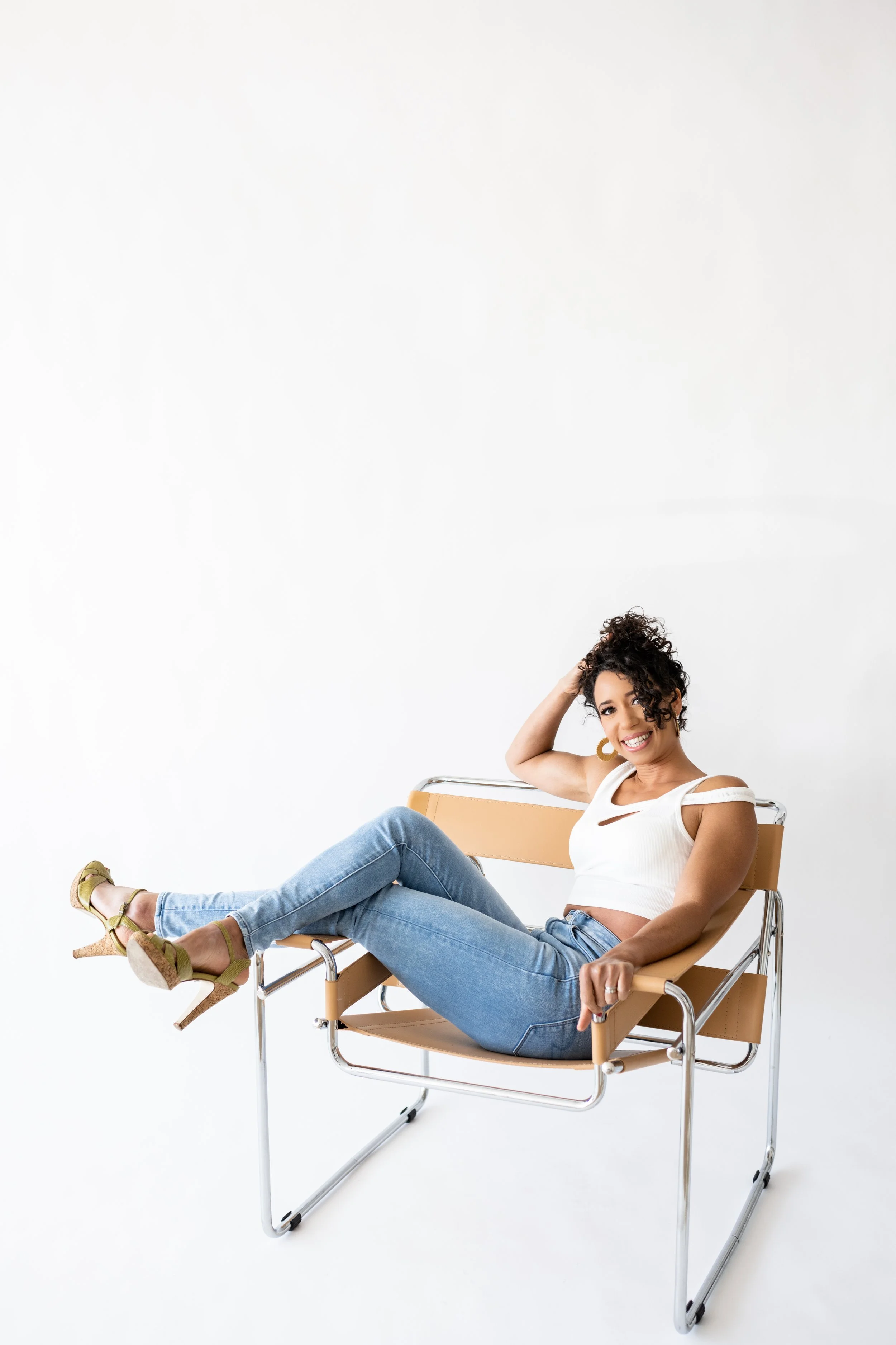 Smiling woman in white crop top and blue jeans sitting on a modern beige chair with a chrome frame against a plain white background.