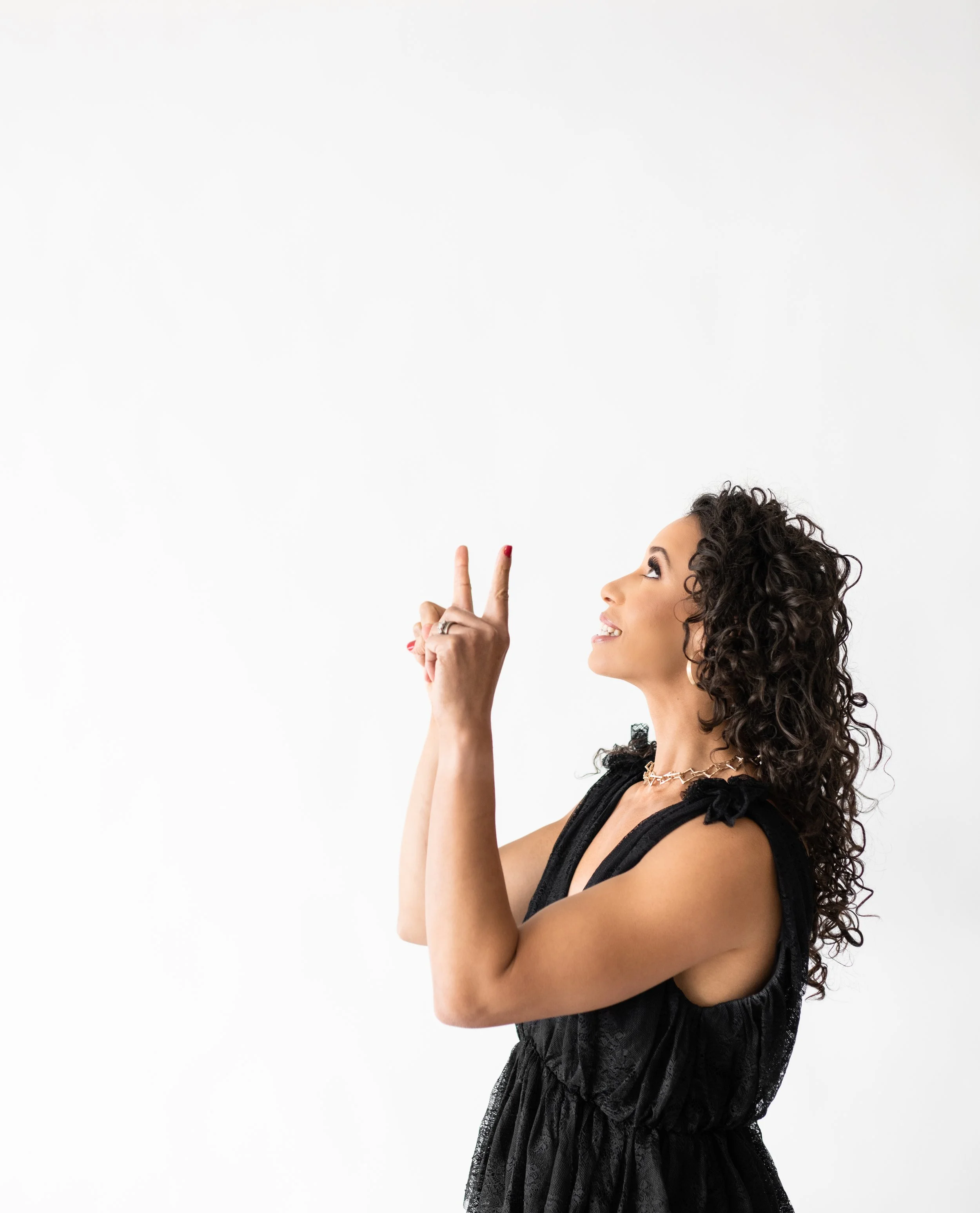 A woman with curly hair wearing a black dress making a peace sign with her right hand and looking upwards.