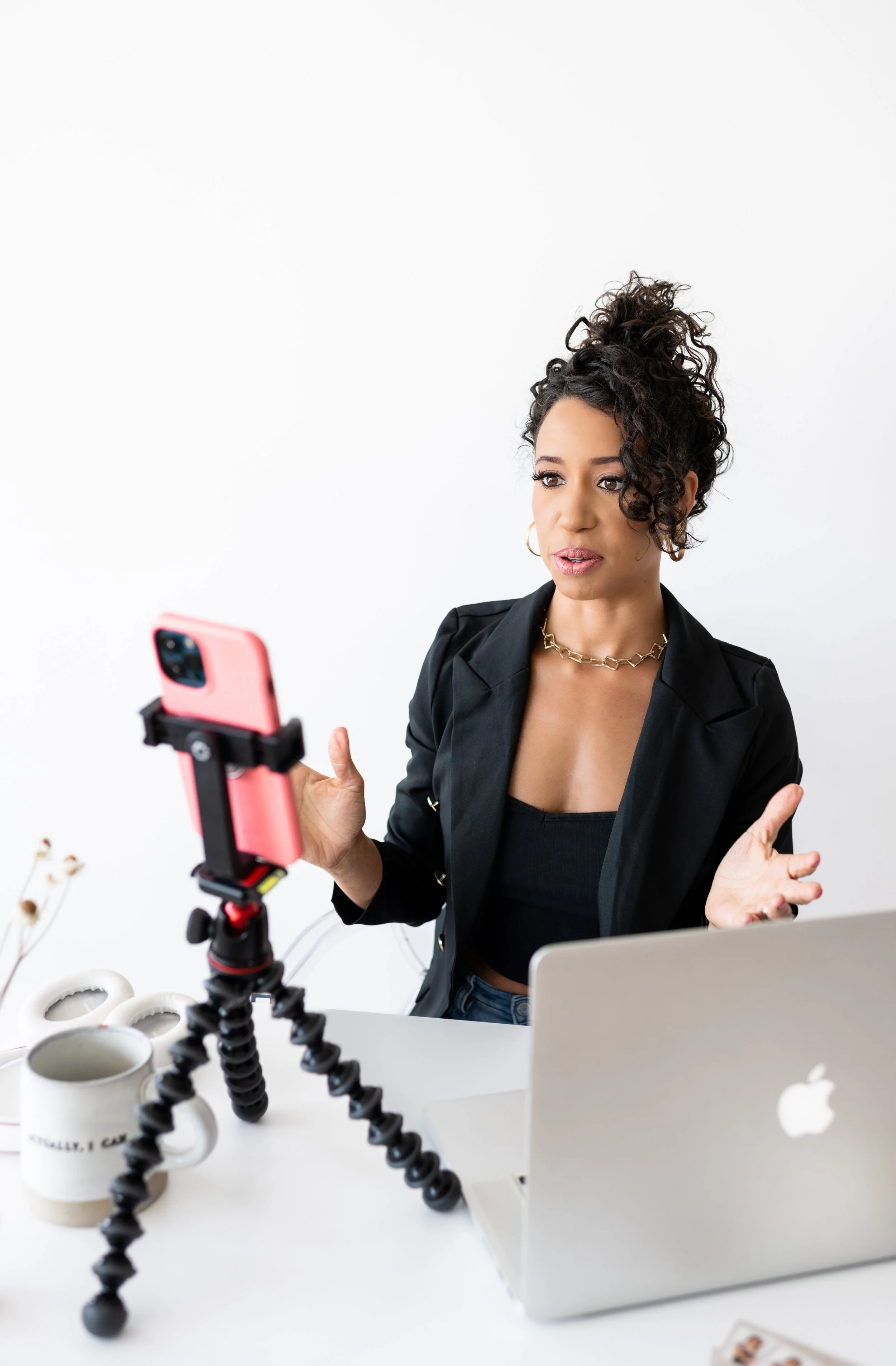 A woman with curly dark hair, wearing a black blazer and gold jewelry, is sitting at a white desk with a MacBook and a camera on a flexible tripod. She appears to be explaining or talking during a video recording.