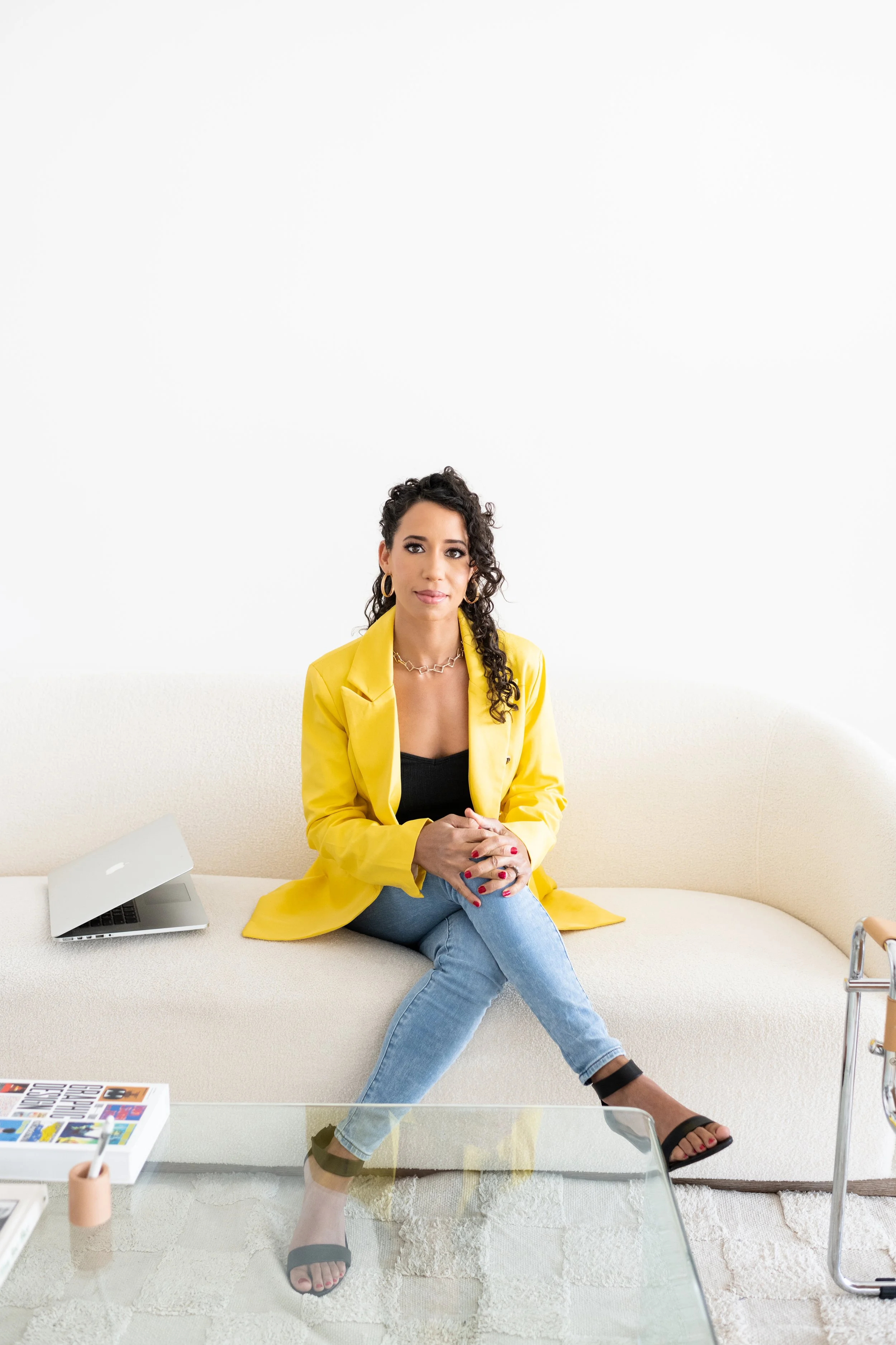 A woman sitting on a cream sofa with a yellow blazer, black top, jeans, and black sandals, with a laptop and colorful magazine in a bright, minimalist room.
