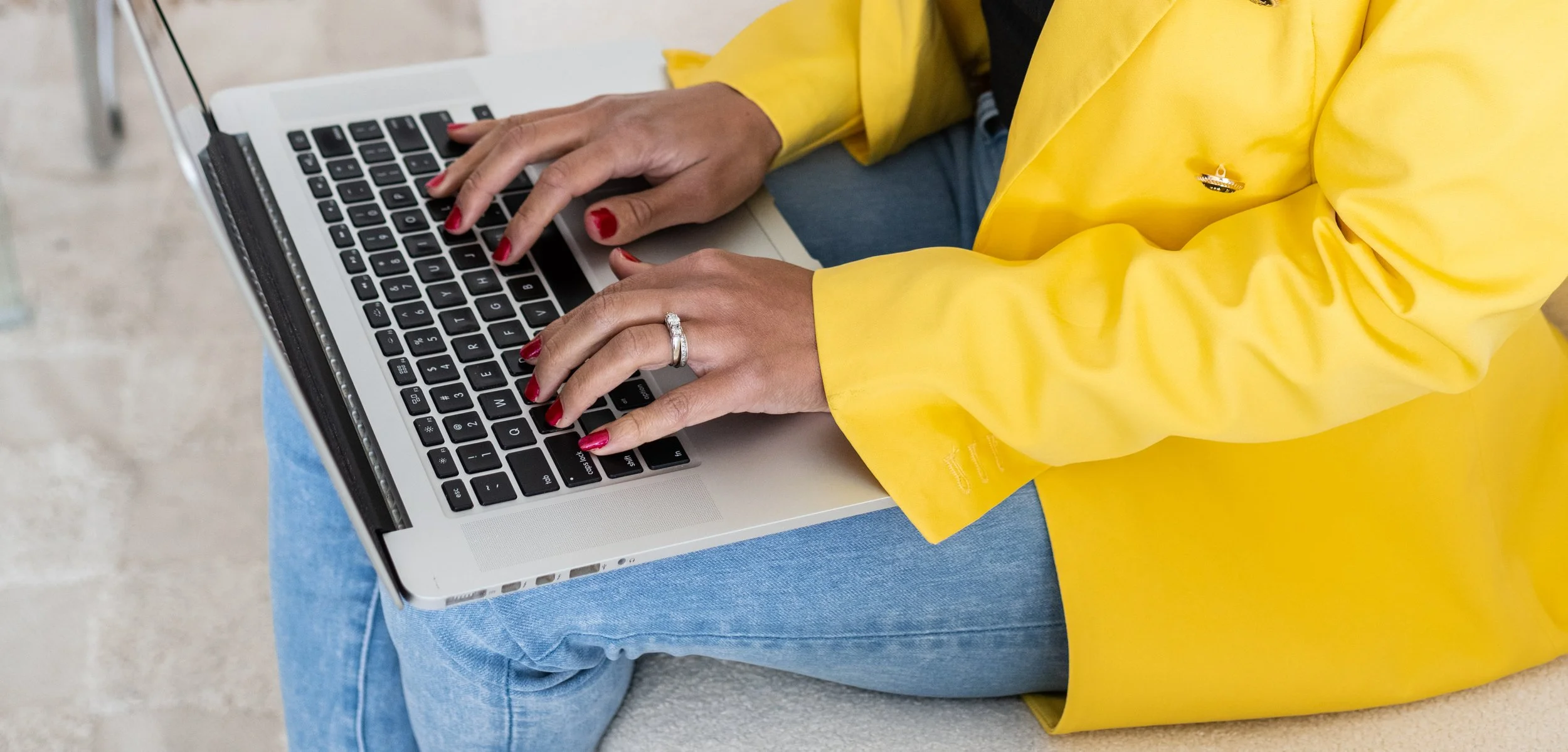 Person wearing a yellow jacket typing on a silver laptop, with red painted nails and a wedding ring, seated on a bench with their legs crossed.