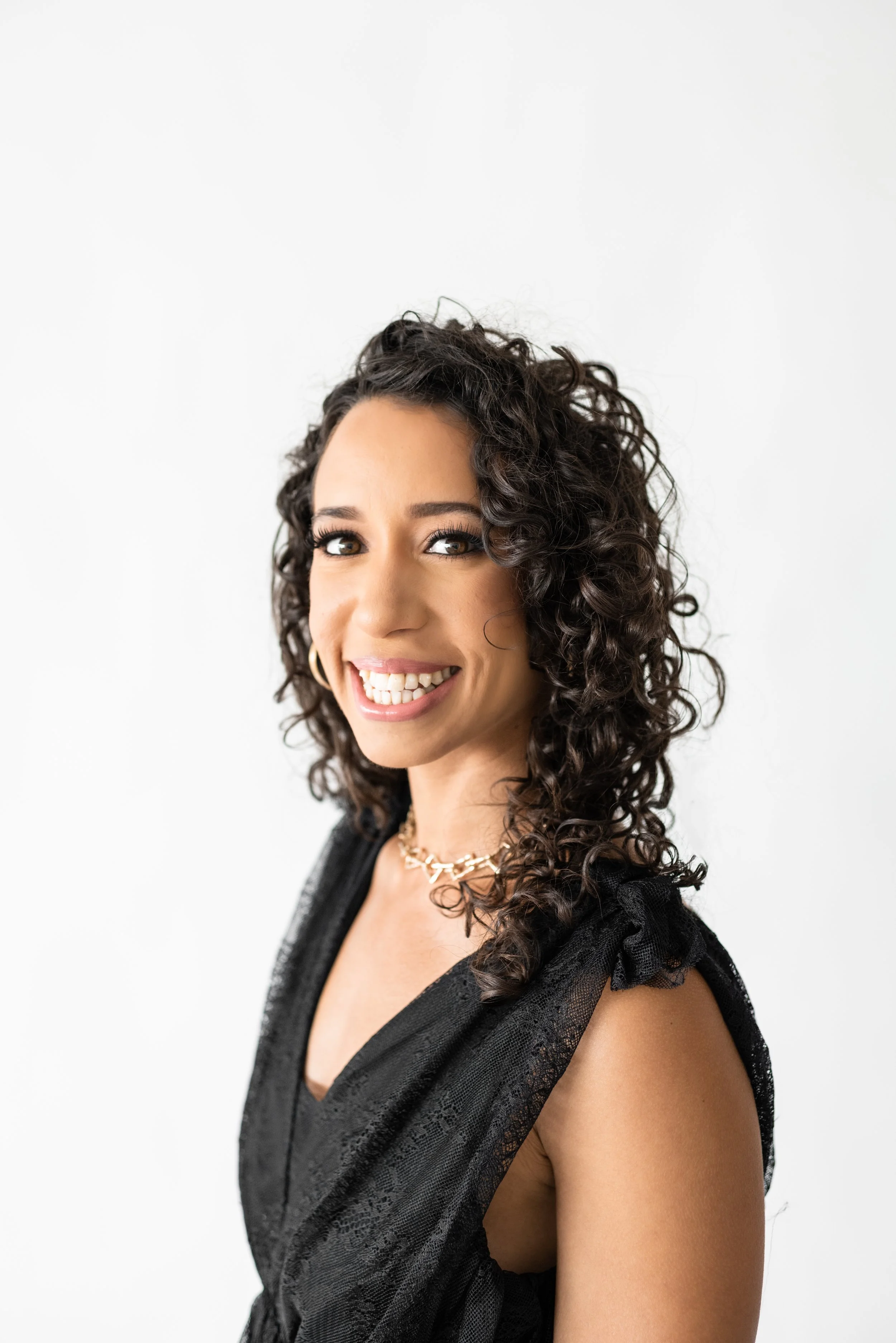 A smiling woman with dark, curly hair wearing a black sleeveless dress and a gold necklace.
