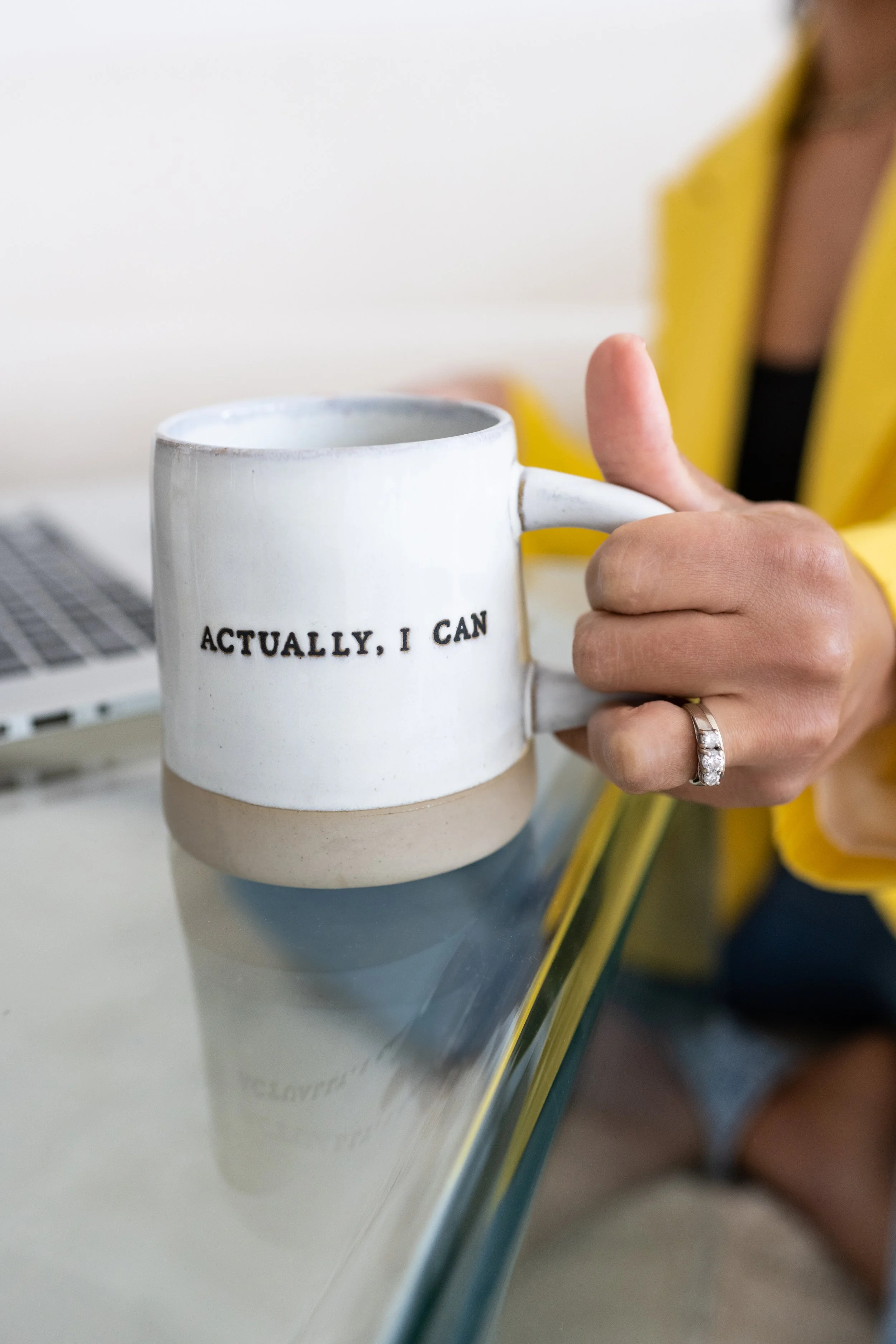 Person holding a white mug with the phrase 'ACTUALLY, I CAN' written on it, sitting at a desk with a laptop.