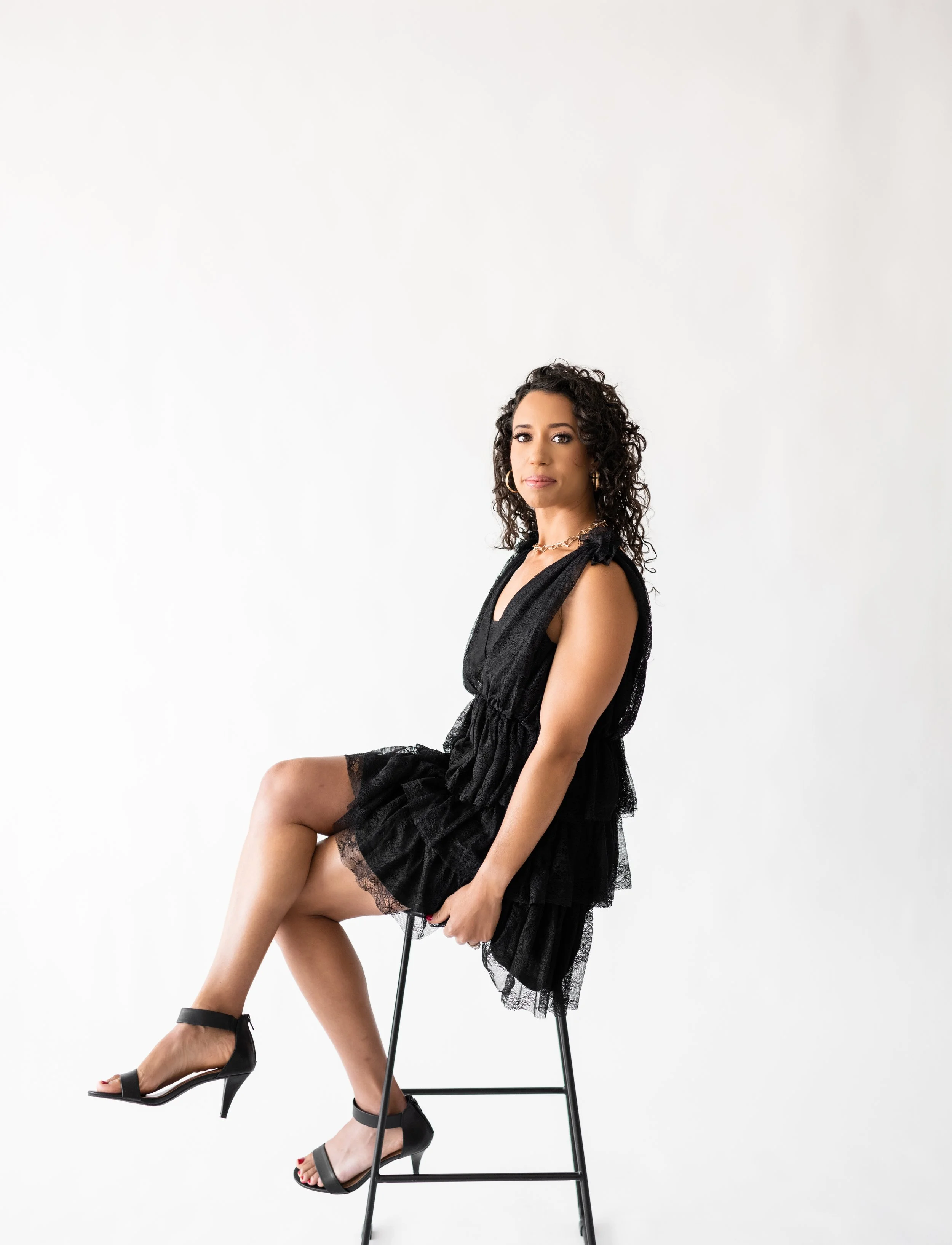 A woman with curly hair seated on a black stool in front of a plain white background, wearing a black dress and black high-heeled shoes.
