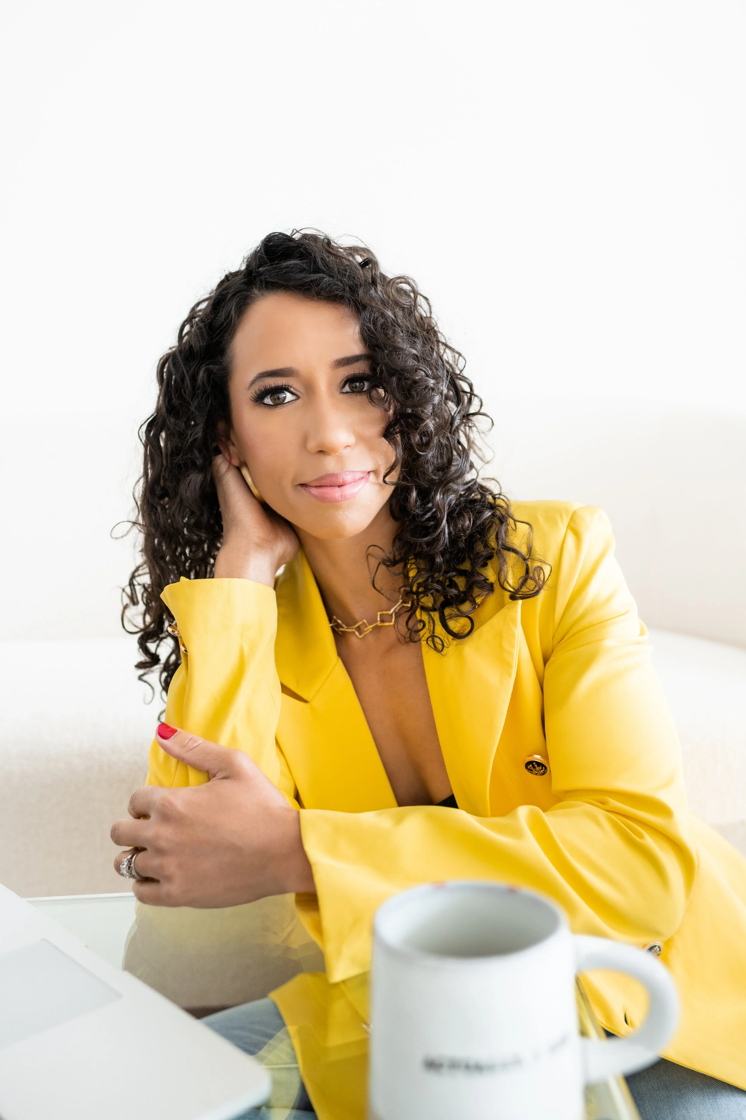 A woman with curly hair wearing a yellow blazer sitting at a table with a mug and a laptop, looking at the camera.