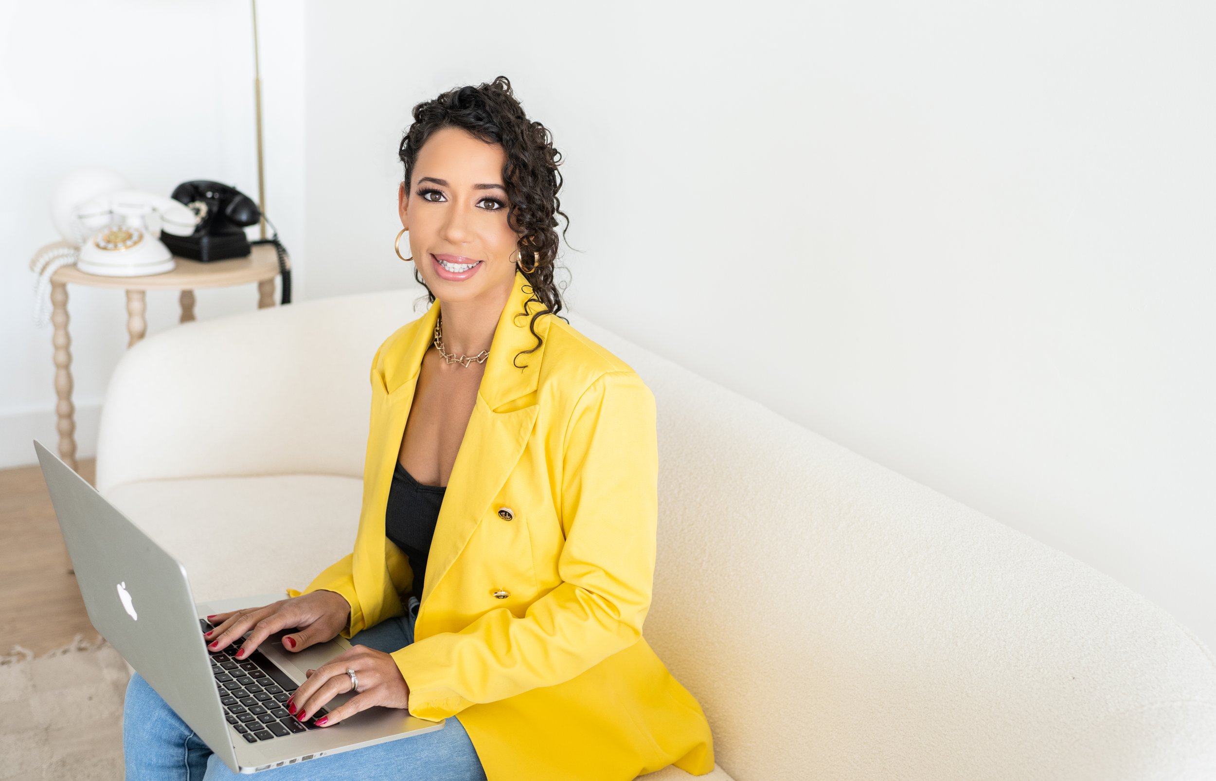 A woman with curly dark hair, wearing a yellow blazer and black top, sitting on a cream-colored sofa using a silver MacBook laptop.