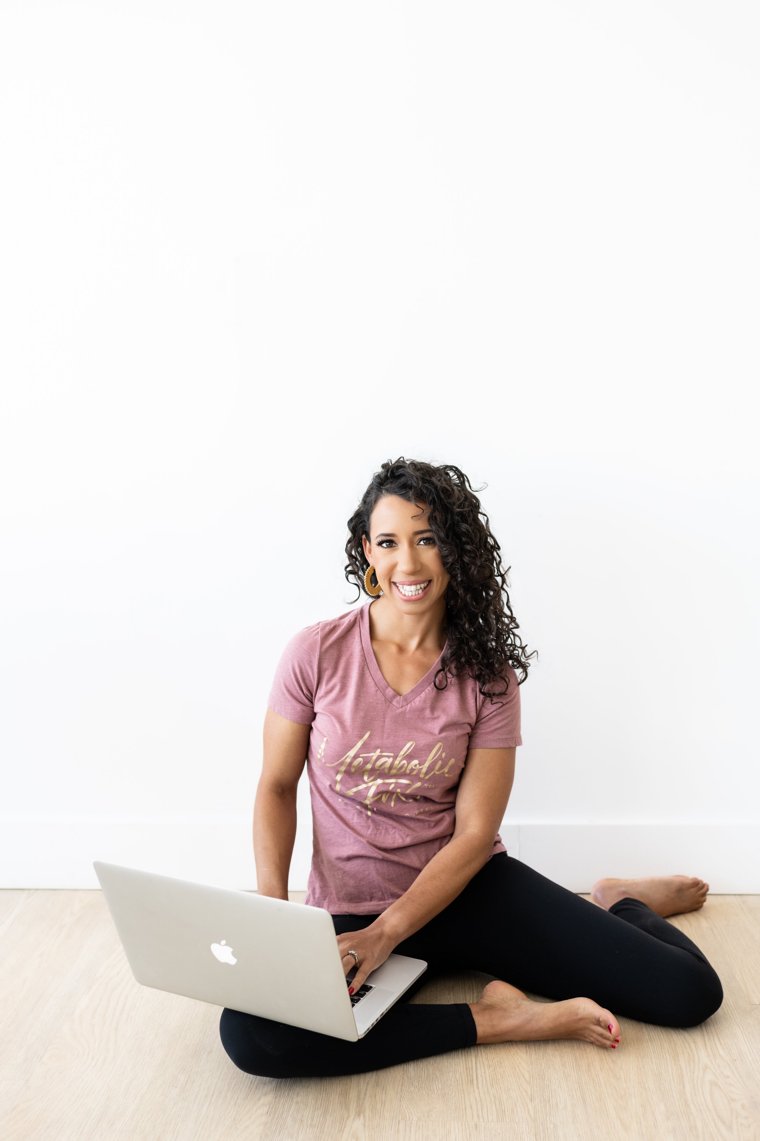 A woman sitting on the floor with a laptop, smiling at the camera, against a white wall.