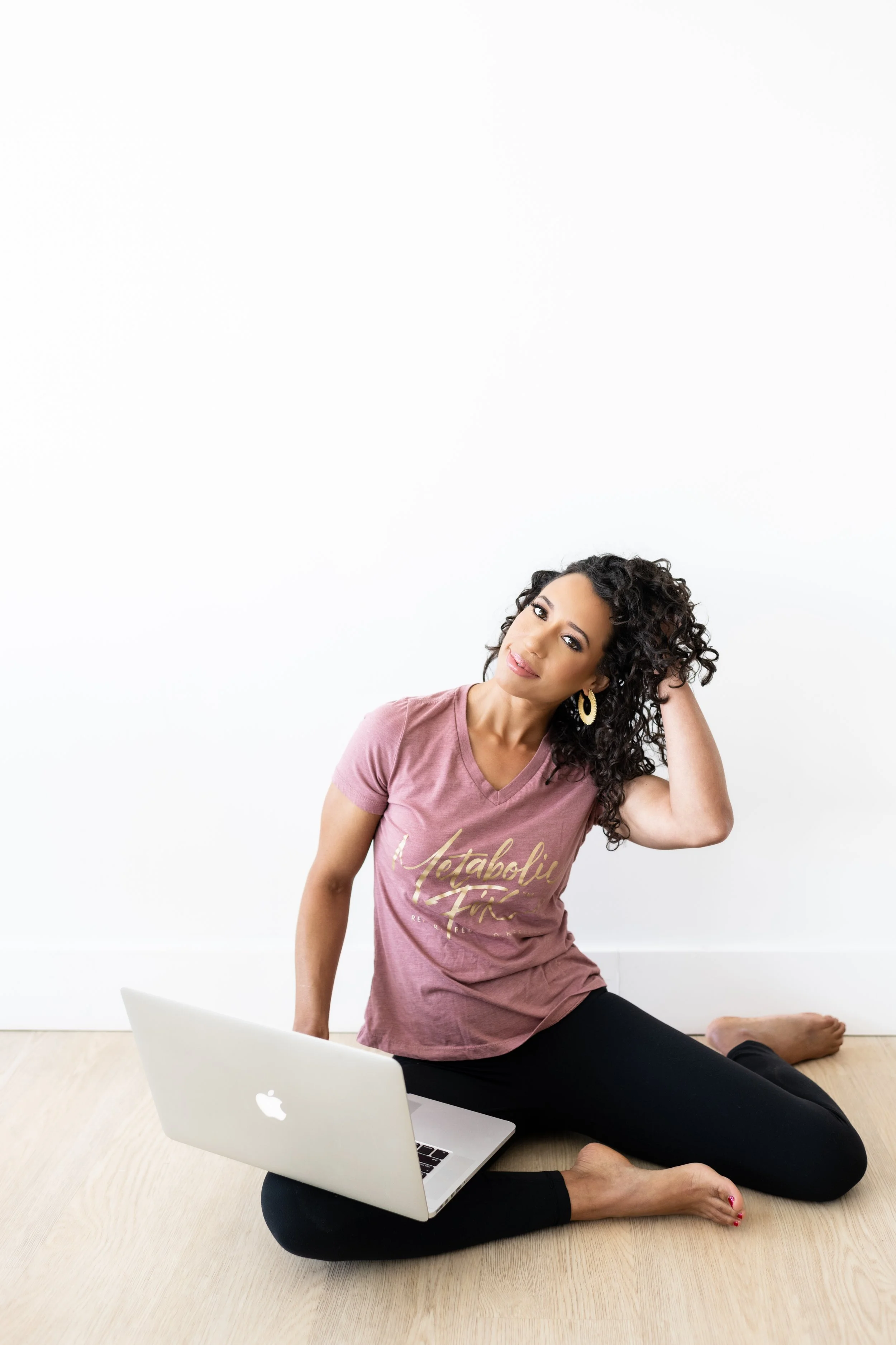 Woman with curly hair sitting on the floor with a laptop, touching her head, against a white wall, wearing a pink shirt and black leggings.