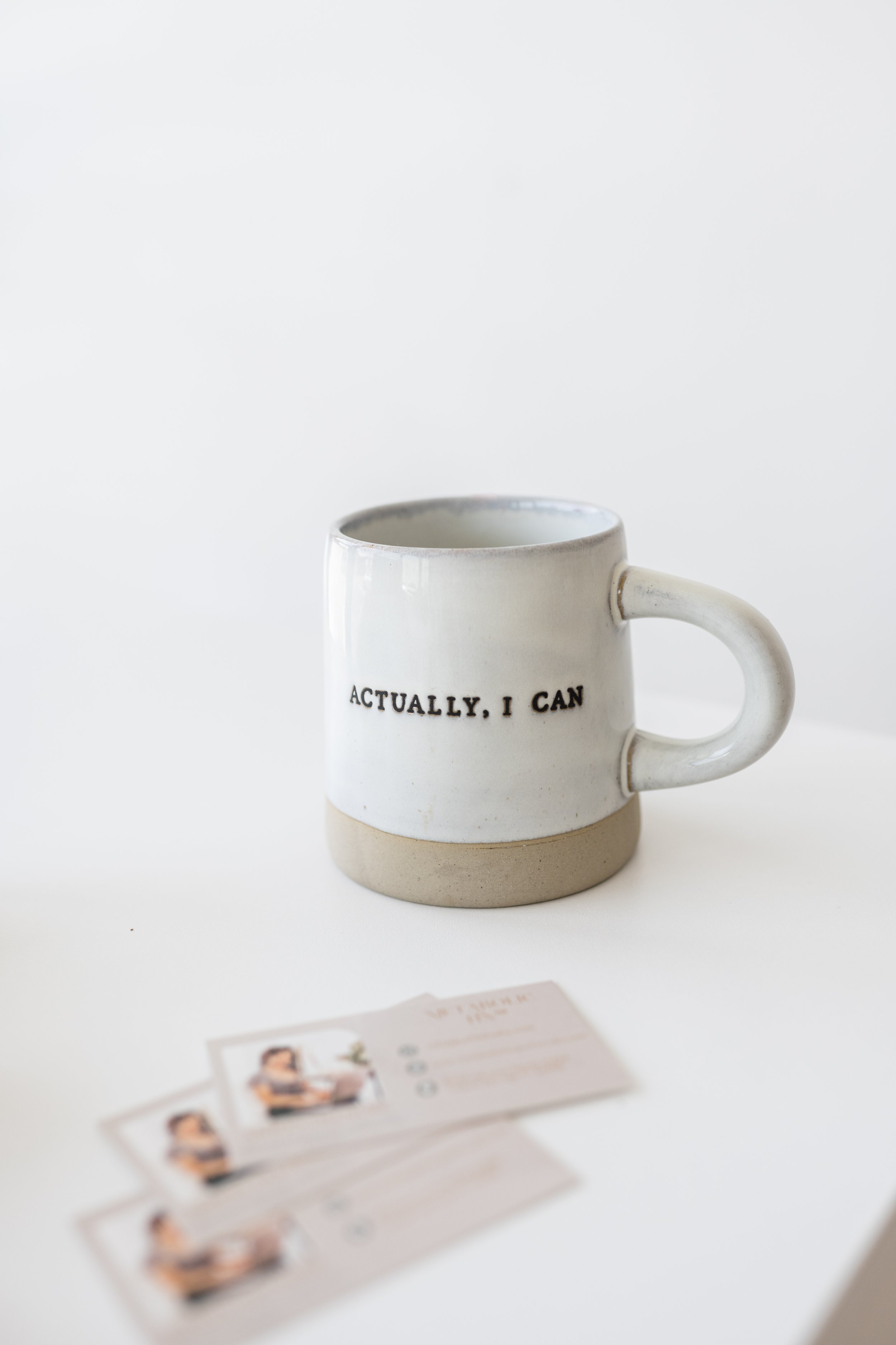 White ceramic mug with black text that says "ACTUALLY, I CAN" and a beige bottom, situated on a white surface with blurred business cards nearby.