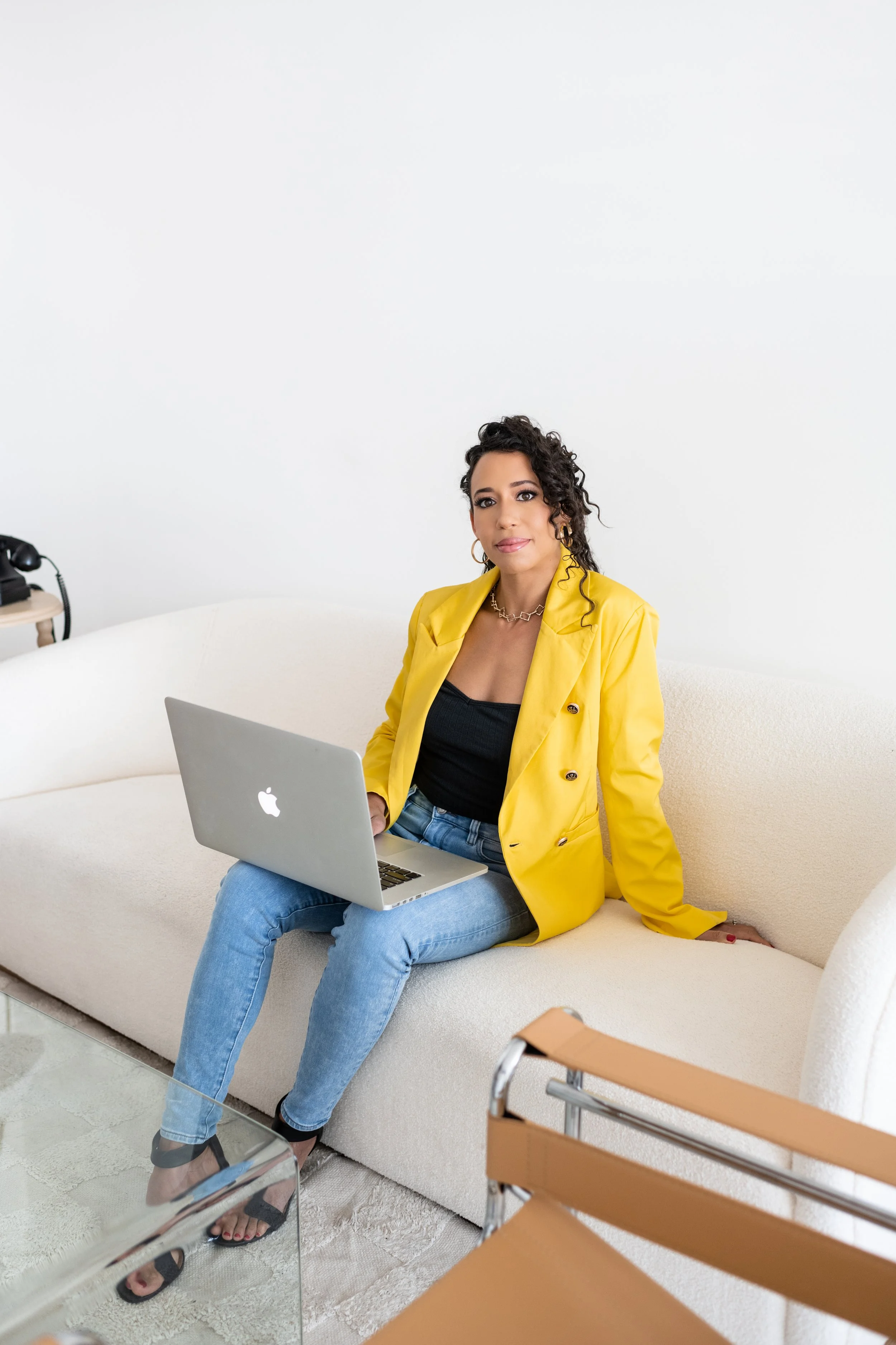 A woman with curly dark hair and gold jewelry wearing a yellow blazer and jeans sitting on a white sofa, holding a silver MacBook laptop in her lap, in a minimalist room with a white wall.