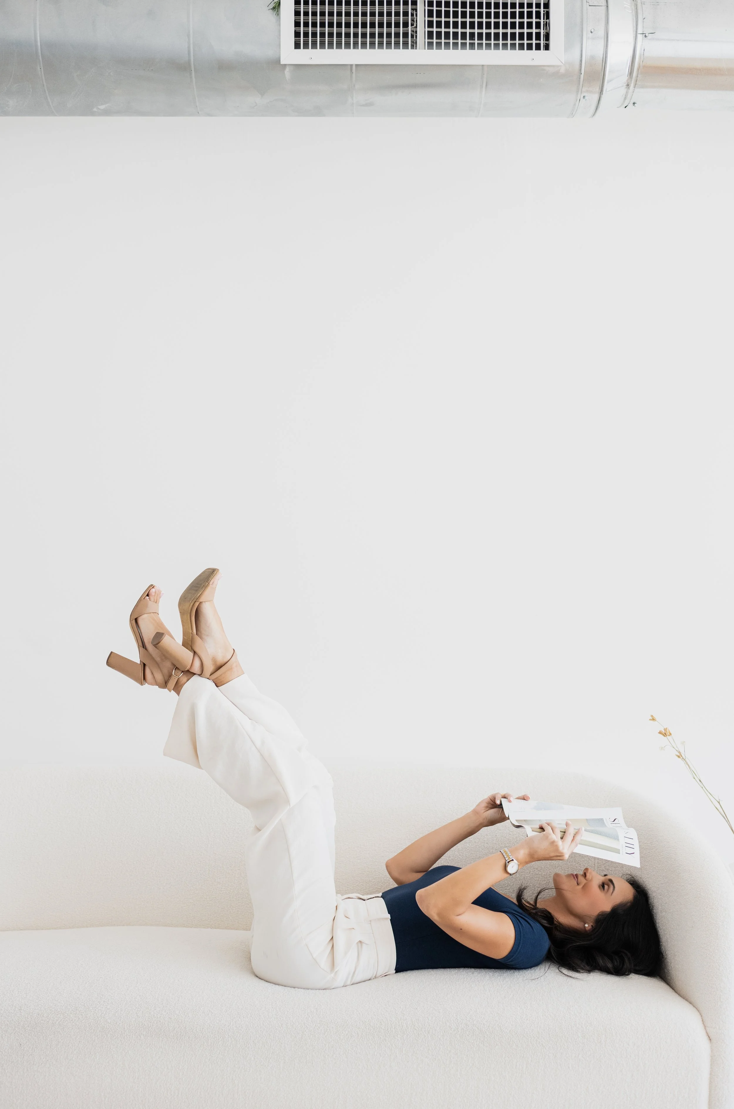 Woman lying on her back on a white sofa, reading a magazine, with her legs raised in the air, wearing beige high heels and a navy blue top.