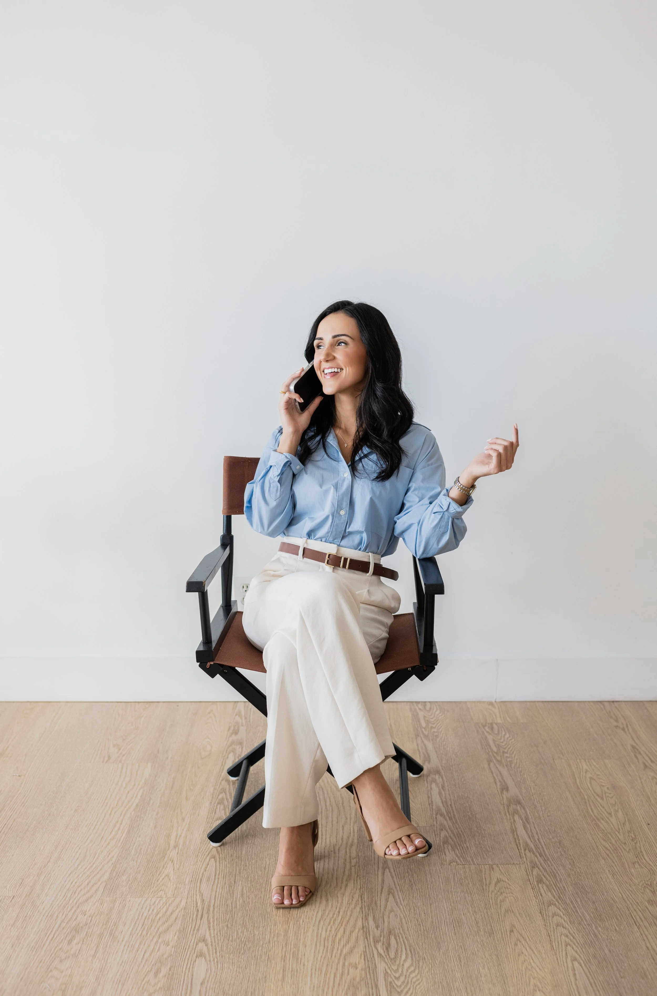 Woman sitting on a director's chair, talking on a phone, smiling, dressed in a blue blouse, beige pants, and tan high heels, against a plain white wall with wooden floor.