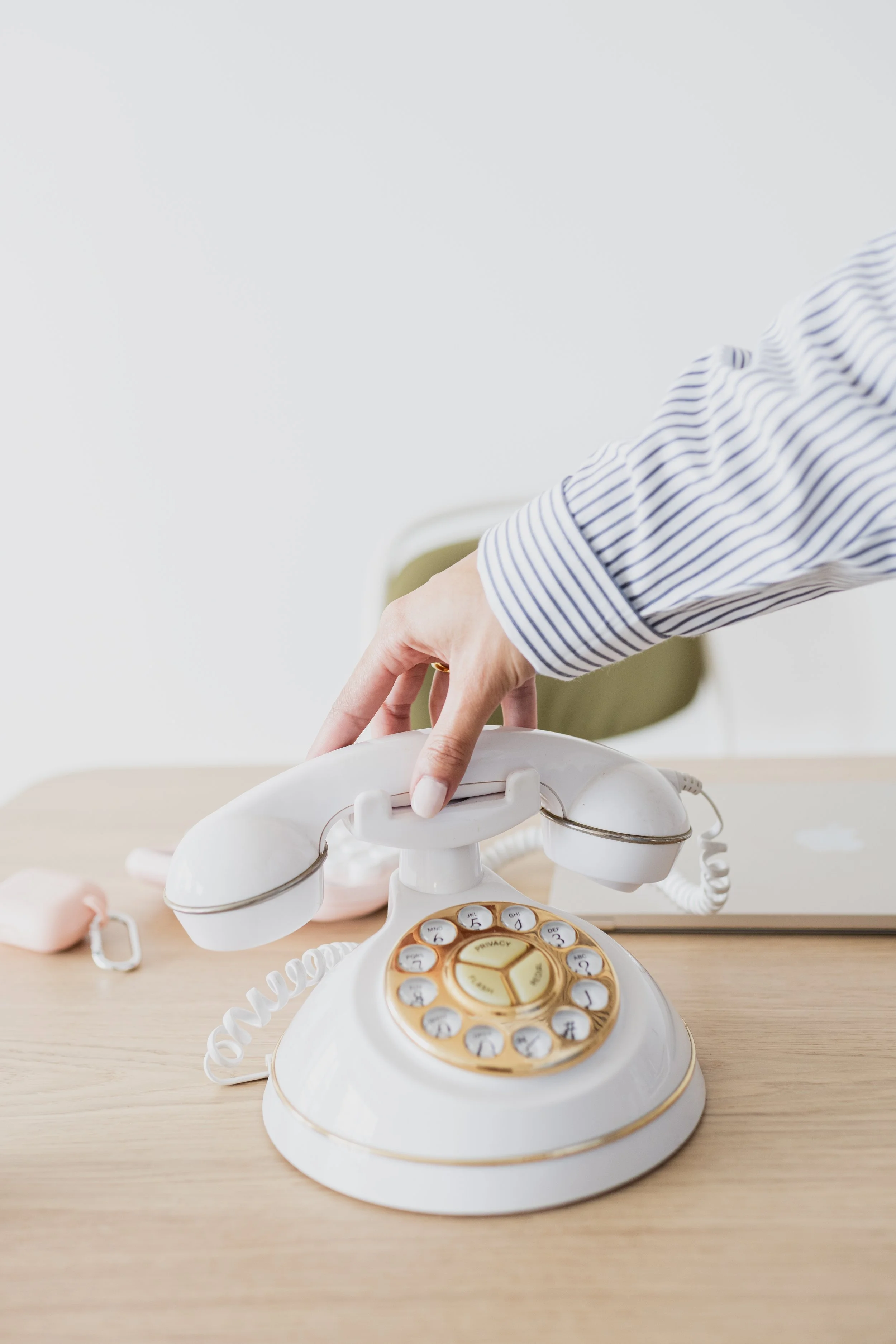 A person in a striped shirt reaching to pick up a vintage white rotary telephone on a wooden desk, with a blurred background that includes a laptop and chair.
