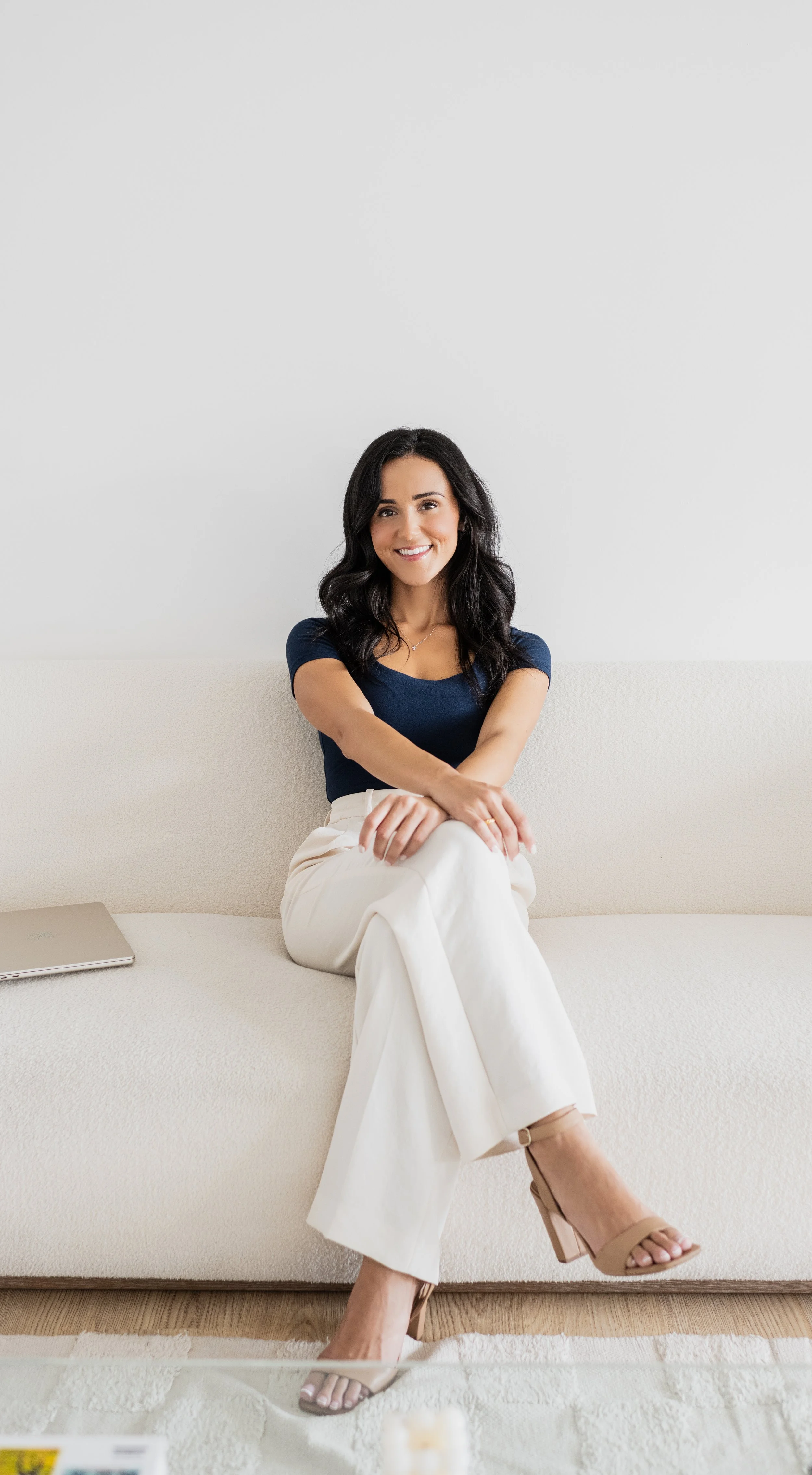 A woman with dark wavy hair, wearing a navy blue top and white trousers, smiling and sitting on a cream-colored sofa with crossed legs, beige heels, and a laptop beside her in a bright room.