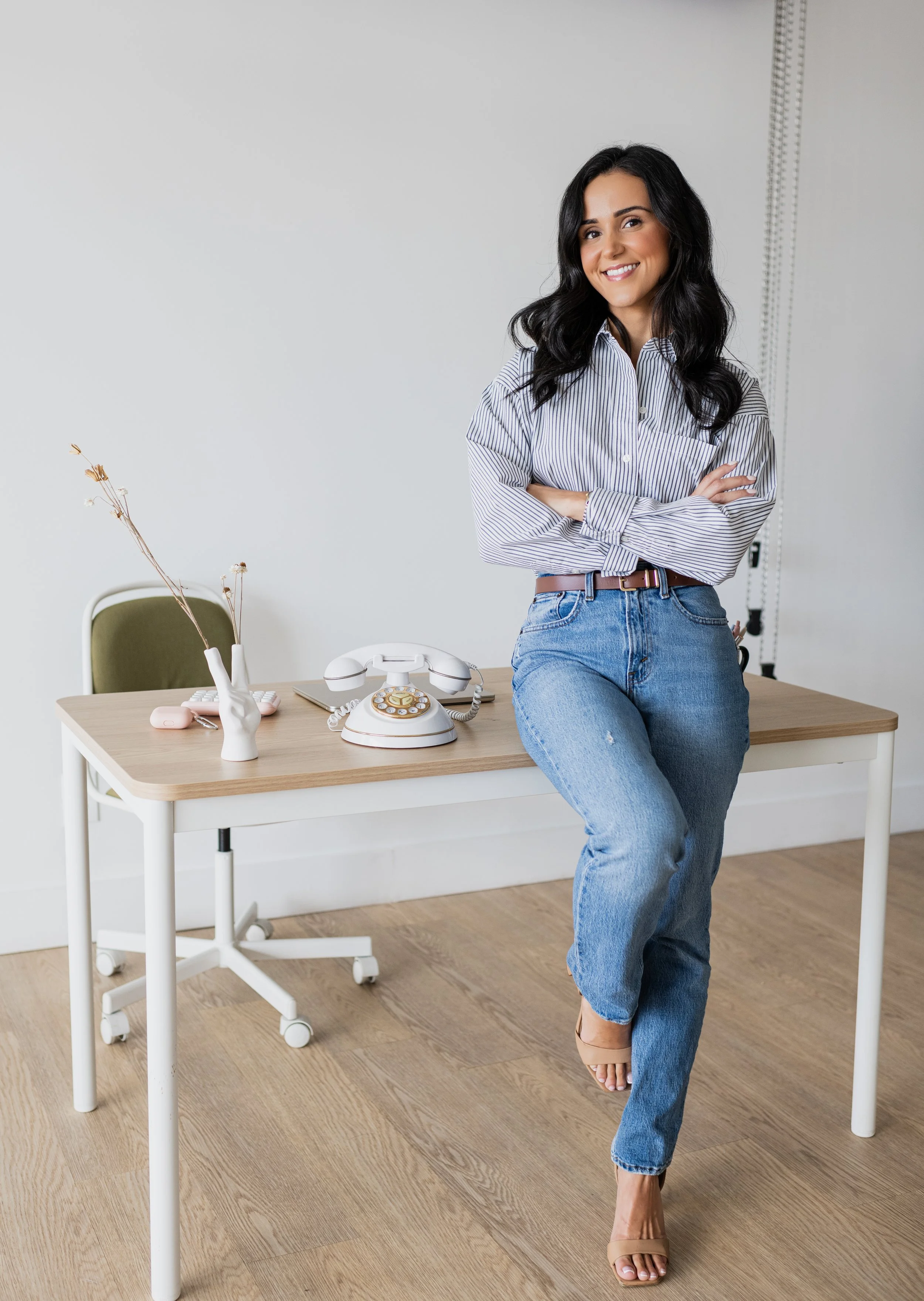 A woman with dark wavy hair, wearing a striped shirt and jeans, standing with crossed arms in an office space, smiling, with a vintage rotary phone and a vase with dried flowers on a desk.