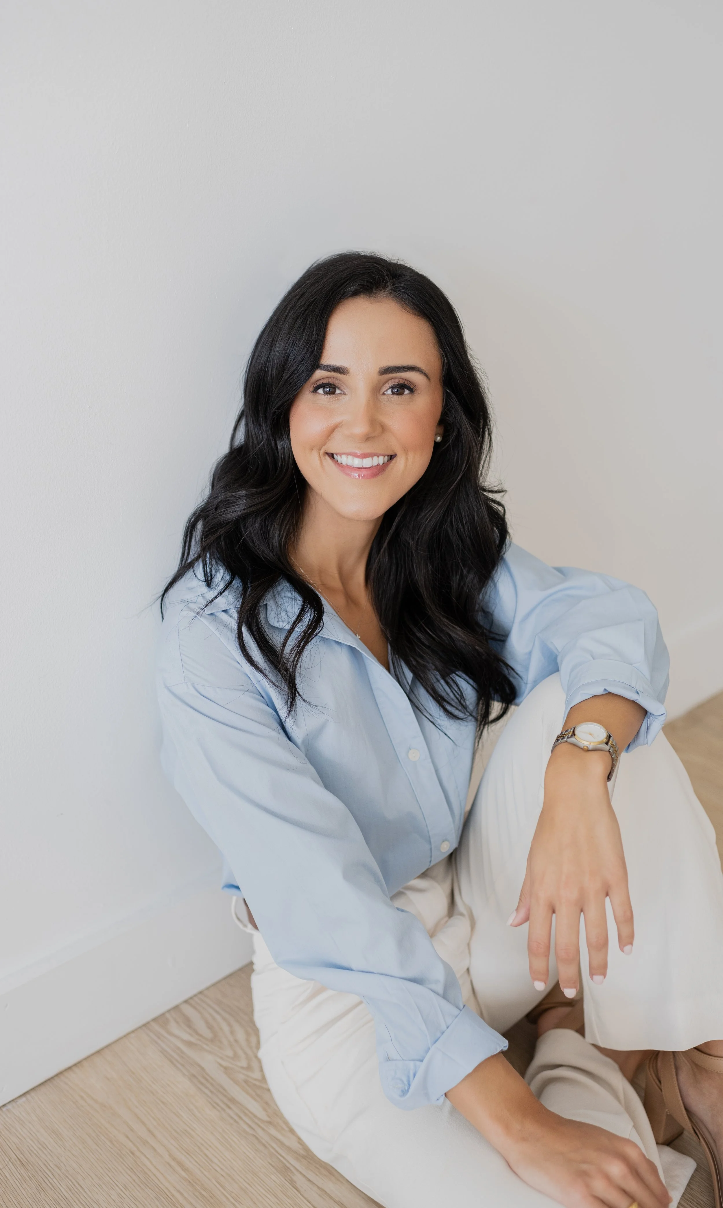 A woman with long black hair, wearing a light blue button-up shirt and cream-colored pants, sitting on a light wooden floor against a white wall, smiling at the camera.