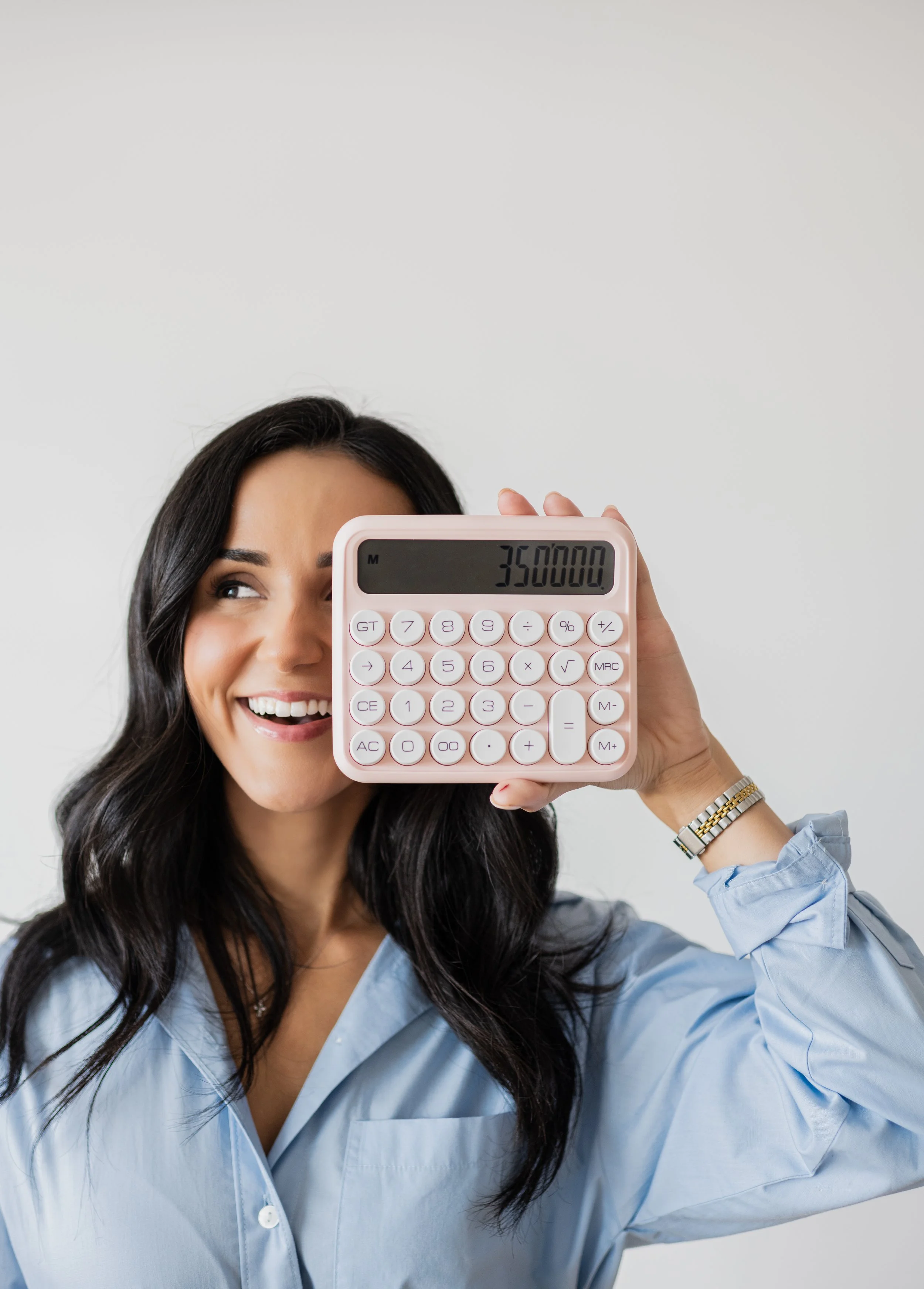 A woman with black hair smiling and holding a pink calculator to her face, showing the number 350000 on the display, wearing a light blue shirt and a silver watch.