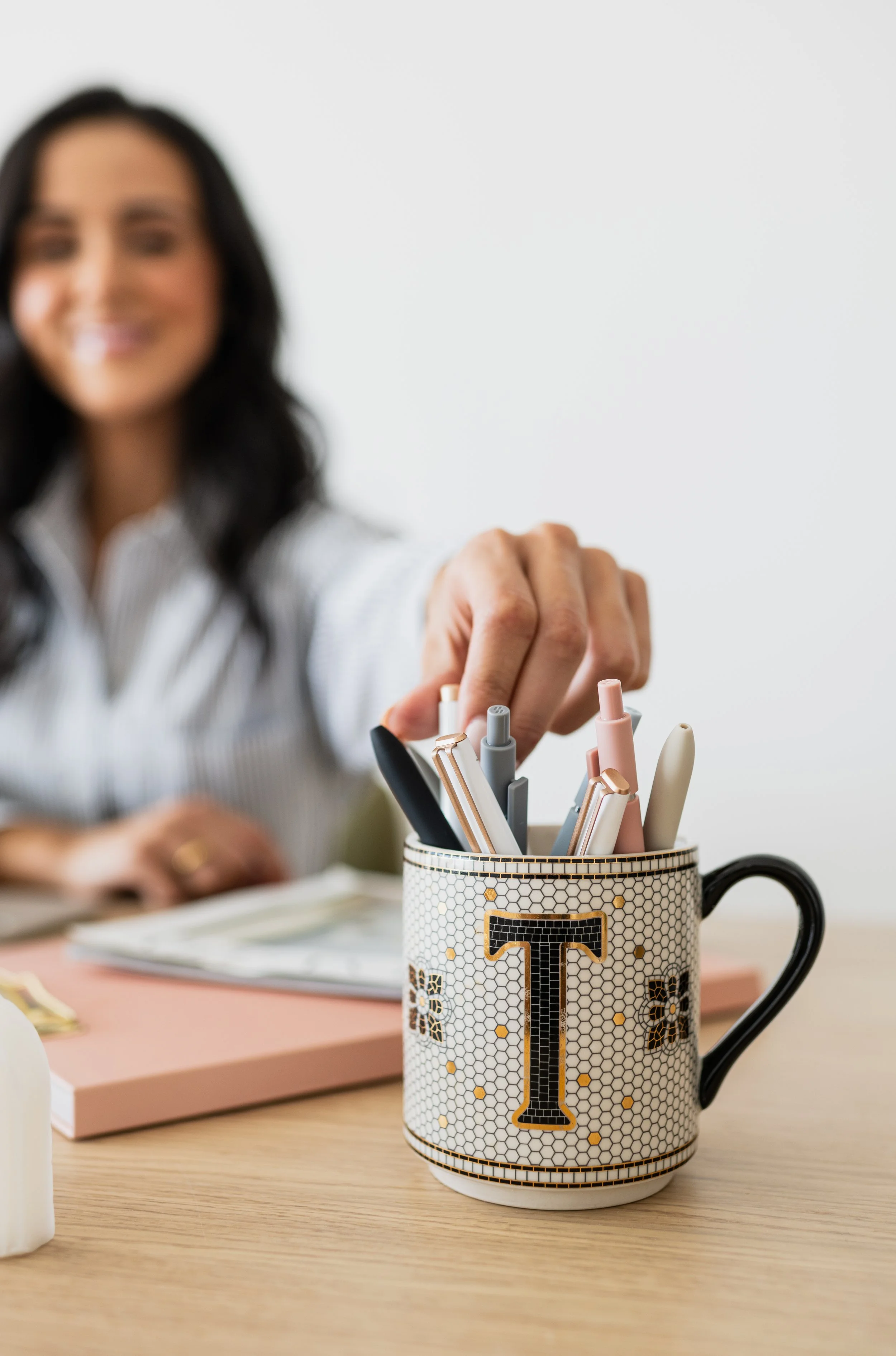 A woman reaching into a mug filled with pens and markers, on a desk with a pink notebook and a closed book behind it, against a plain white background.