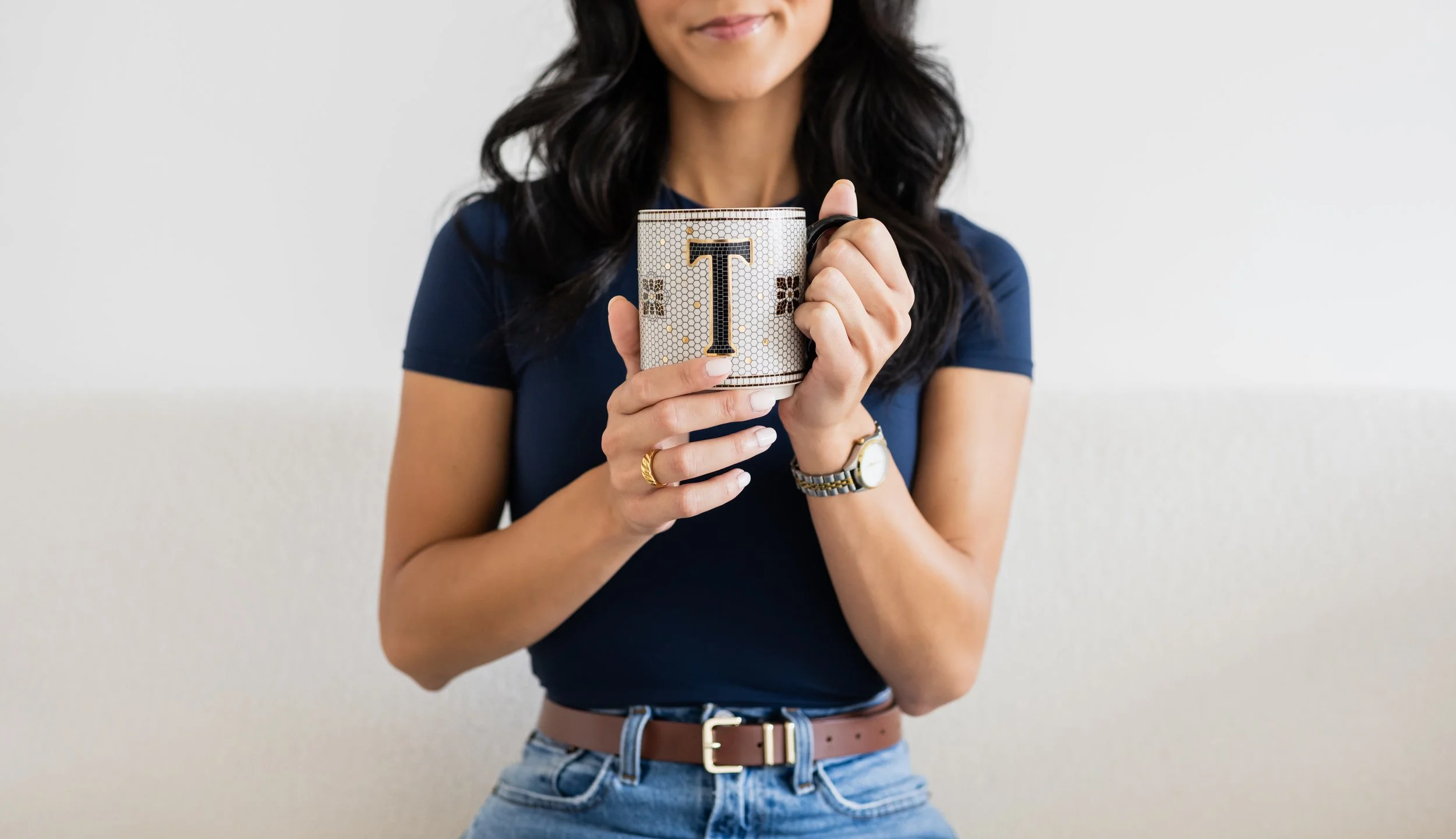 A woman with dark, wavy hair wearing a dark blue t-shirt holds a coffee mug with a large letter 'T' on it. She is standing against a plain light background.