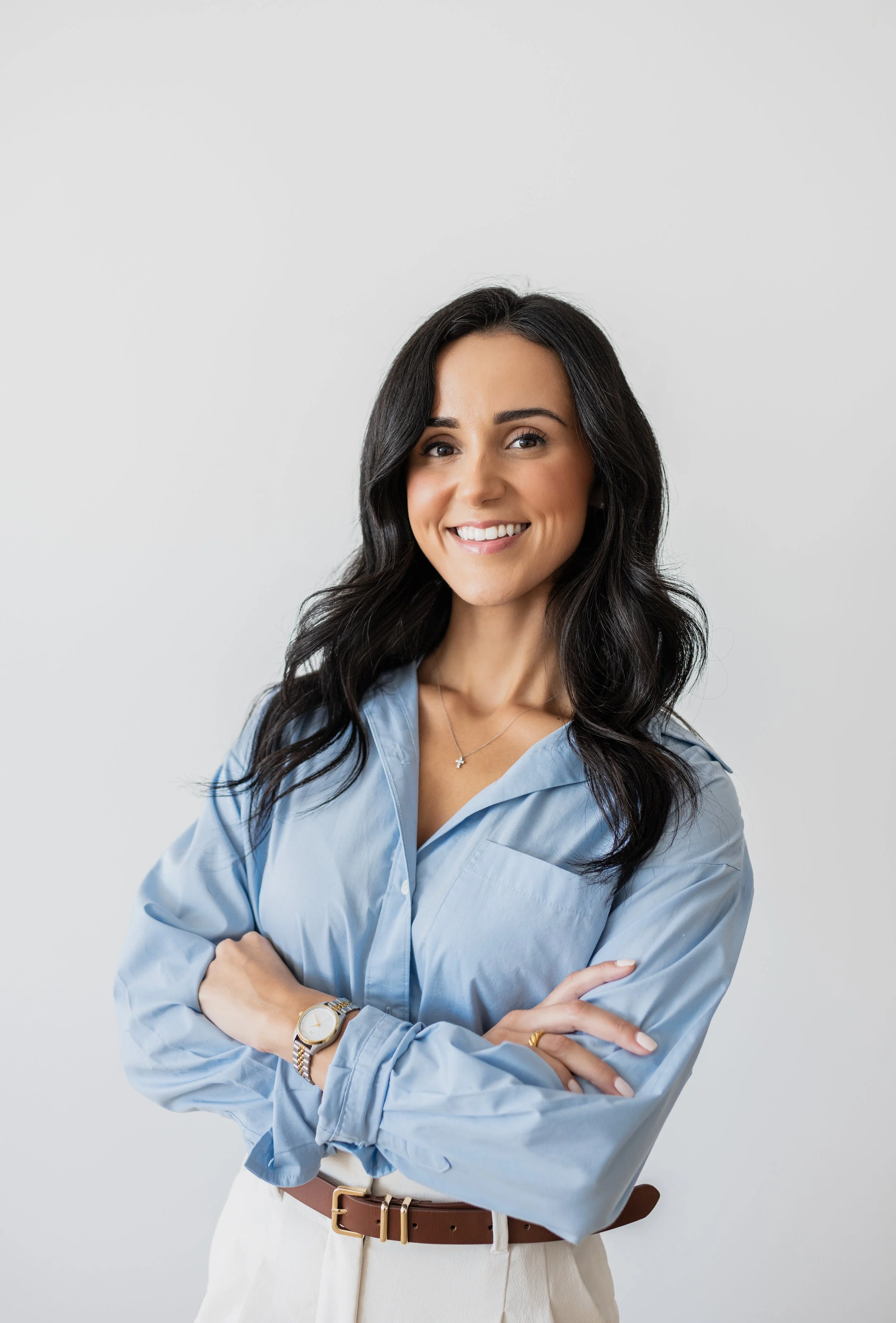 A woman with long dark wavy hair, smiling, wearing a light blue button-up shirt, beige pants, a wristwatch, a necklace, and a ring, standing with her arms crossed against a plain white background.