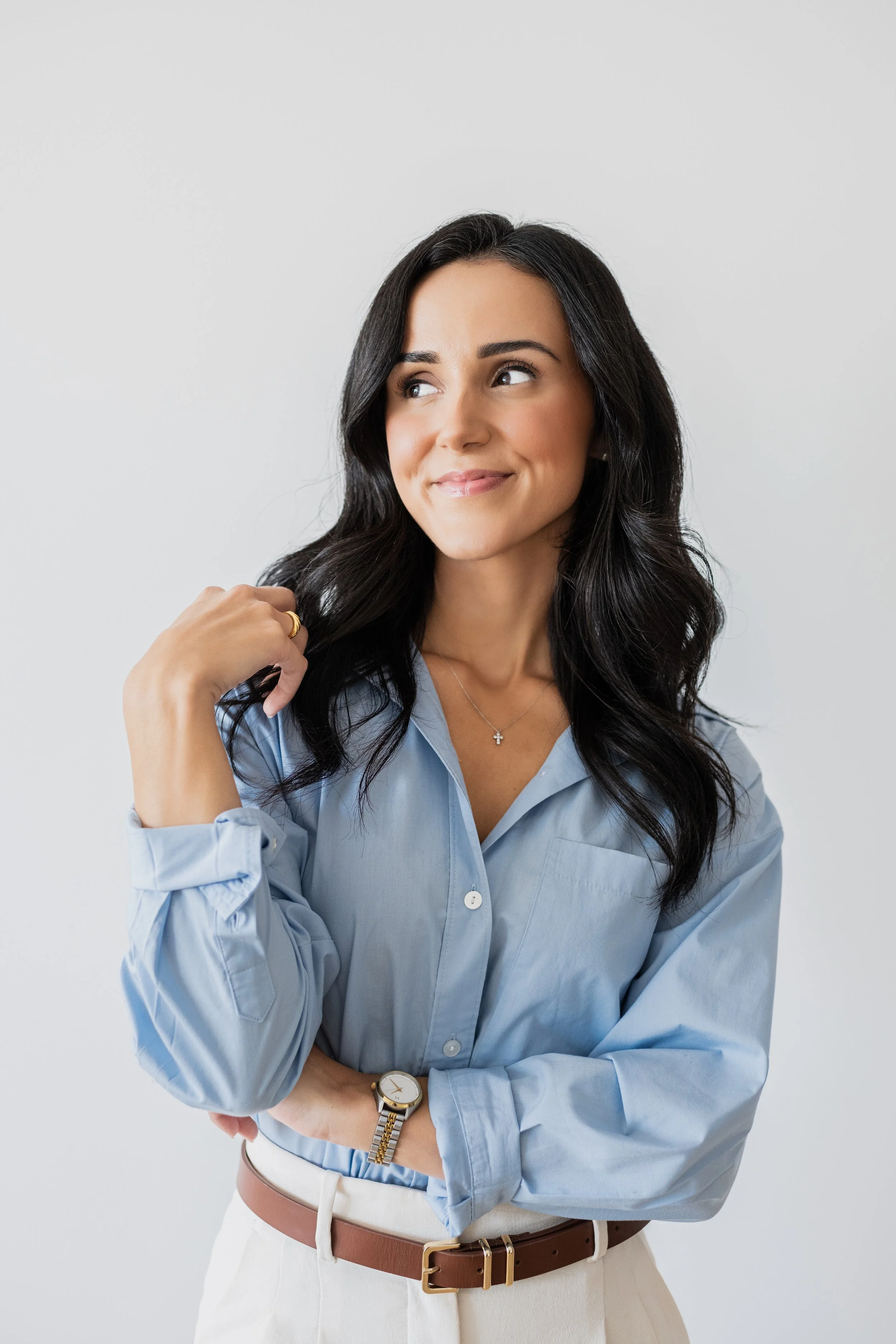 A woman with long black hair wearing a light blue button-up shirt, beige pants, a brown belt, a wristwatch, a necklace, and a ring, smiling and looking to her right against a plain white background.