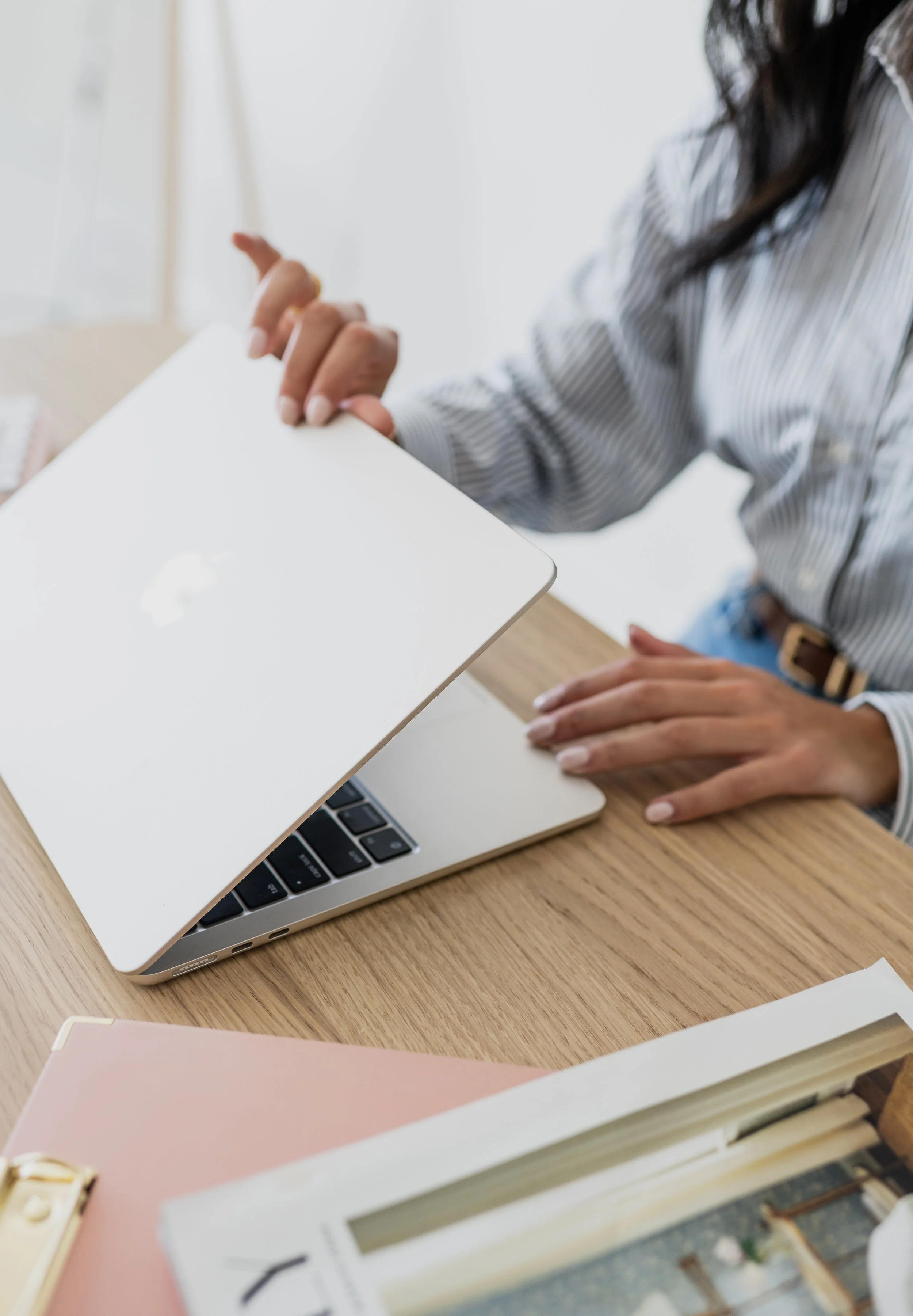 A woman in a striped shirt is at a desk with a silver laptop, a pink notebook, a magazine, and a wooden clipboard, reaching towards the laptop.