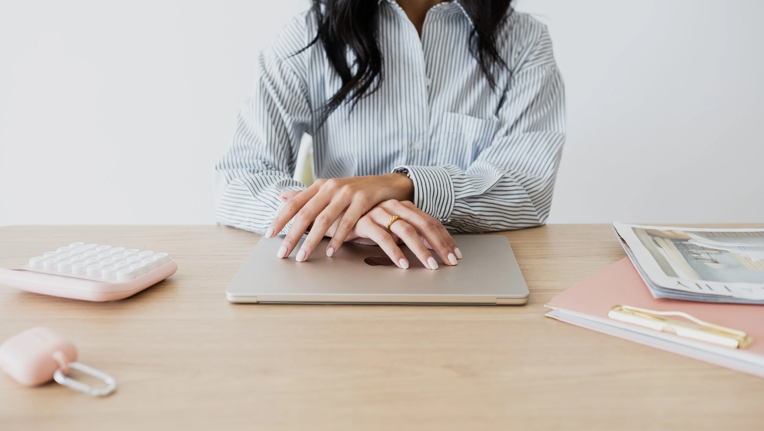 Person with dark hair wearing a striped shirt sitting at a desk with a closed laptop, pink calculator, pink keychain, stacked magazines and pink folder.