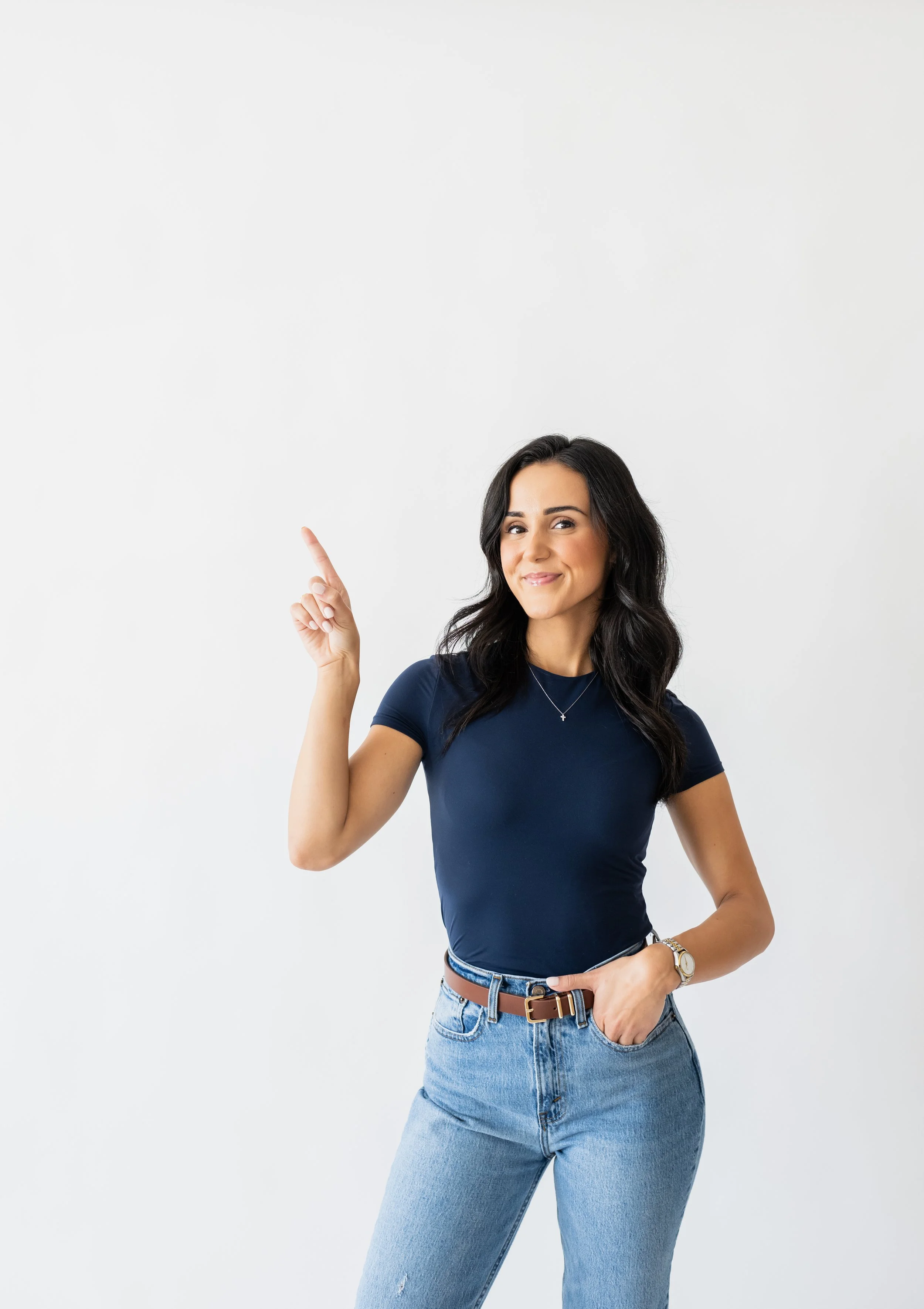 A young woman with long dark hair, wearing a navy blue t-shirt and light blue jeans, pointing upward with her right index finger and smiling, standing against a plain white background.