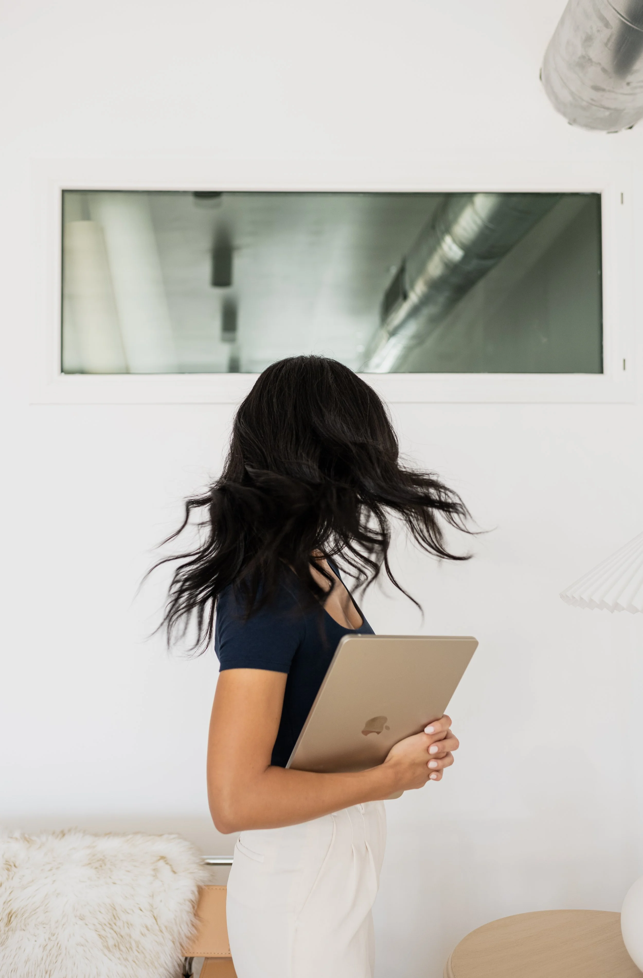 Woman holding a silver MacBook laptop, standing in a modern, minimalistic room with a large window and white walls.