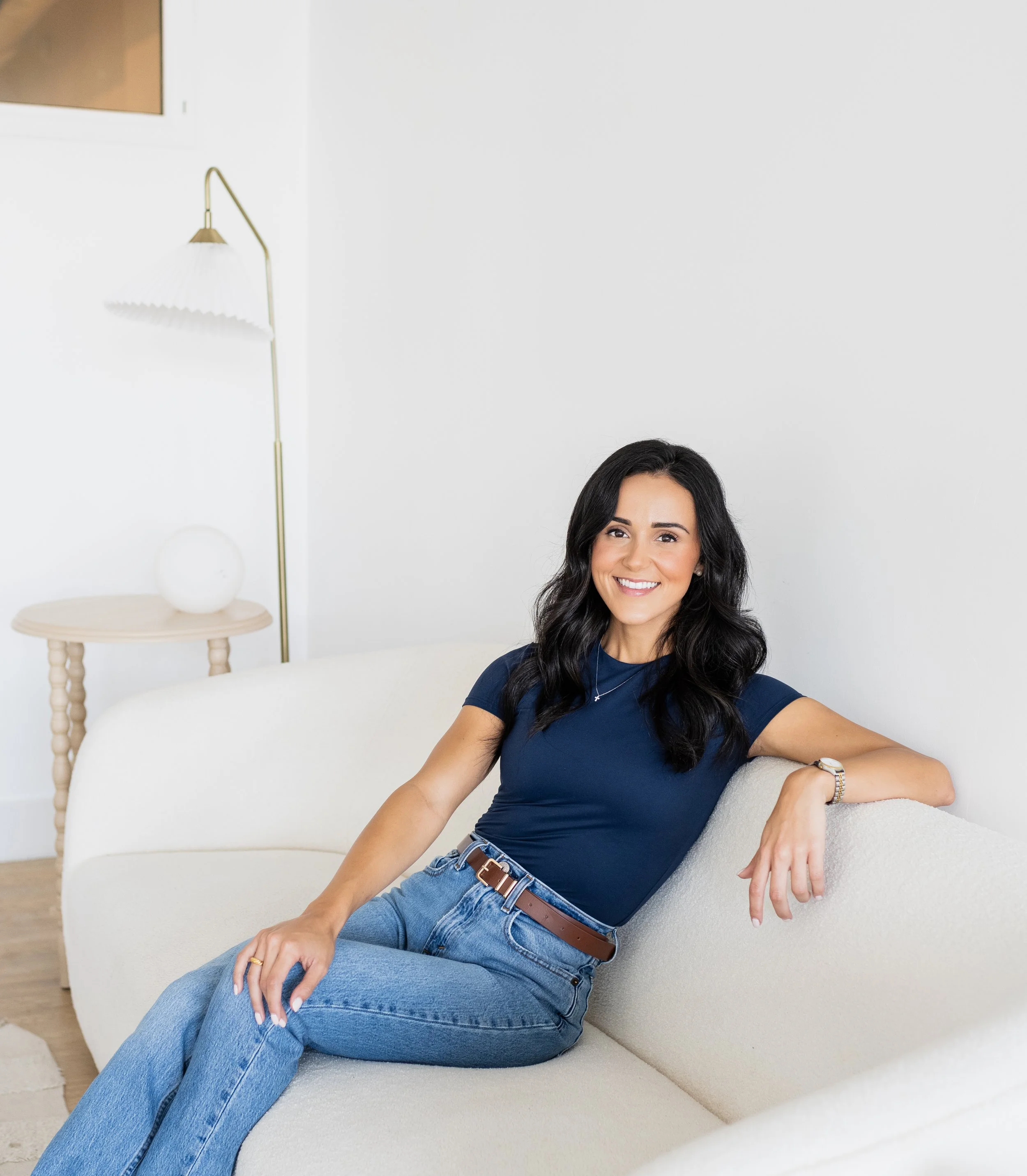A woman sitting on a light-colored sofa in a modern, minimalistic living room, smiling at the camera, wearing a navy blue t-shirt and blue jeans.