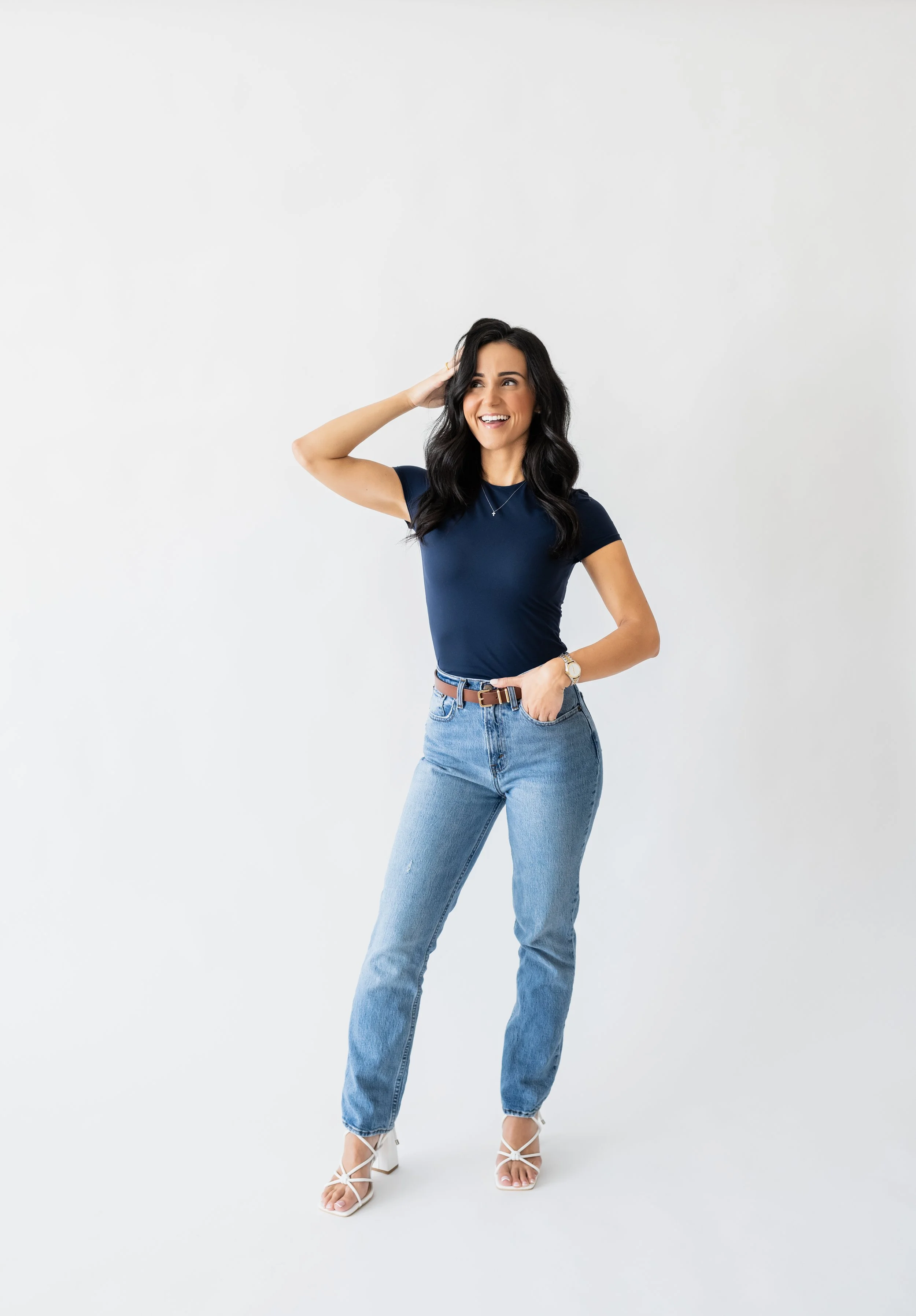 A woman with long dark hair, wearing a navy blue t-shirt, light blue jeans, and white high heels, standing against a white background, smiling and touching her hair.