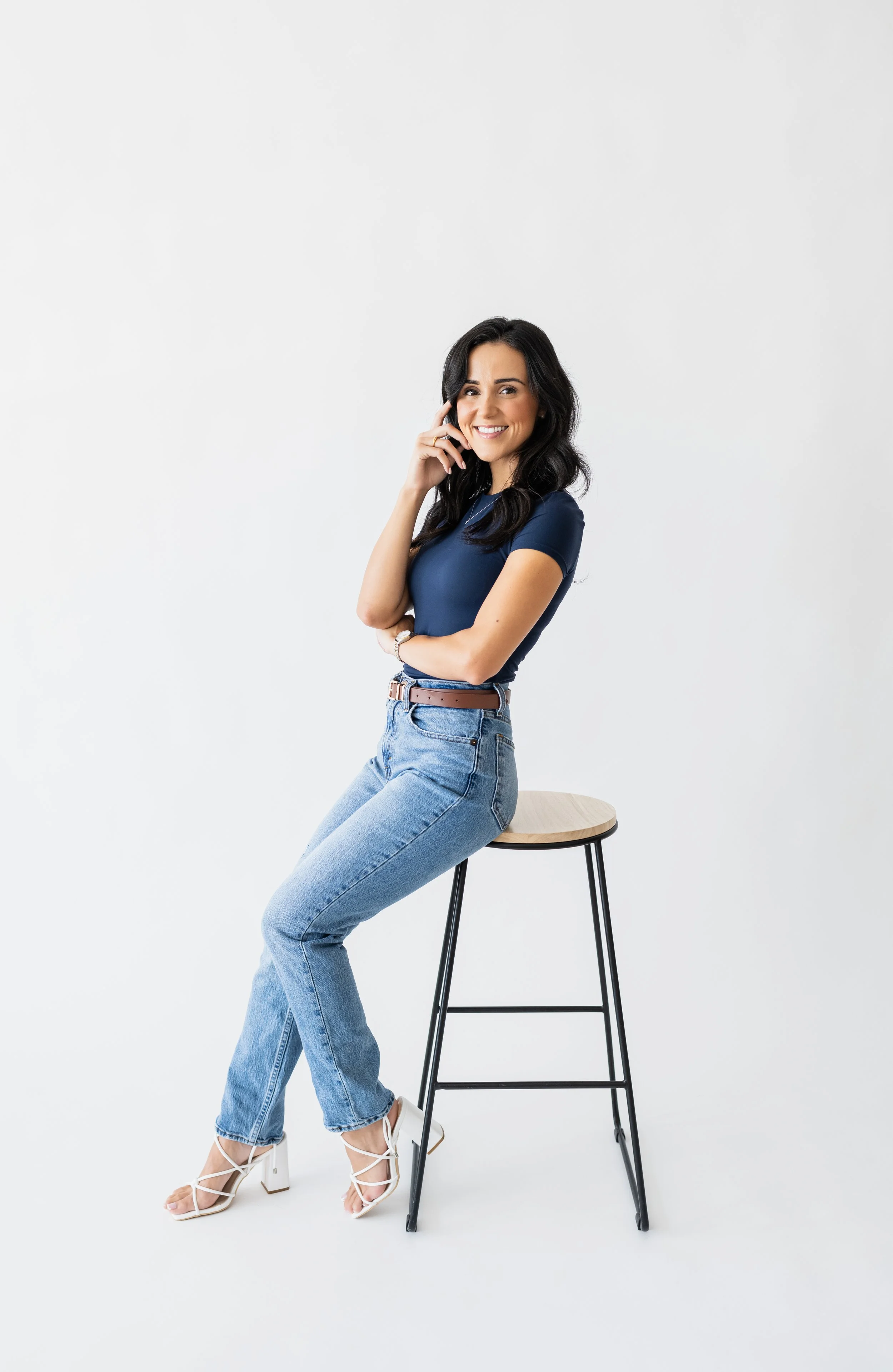 A woman with dark hair sitting on a high stool, smiling, wearing a navy t-shirt, light blue jeans, and white strappy heels, with a plain white background.