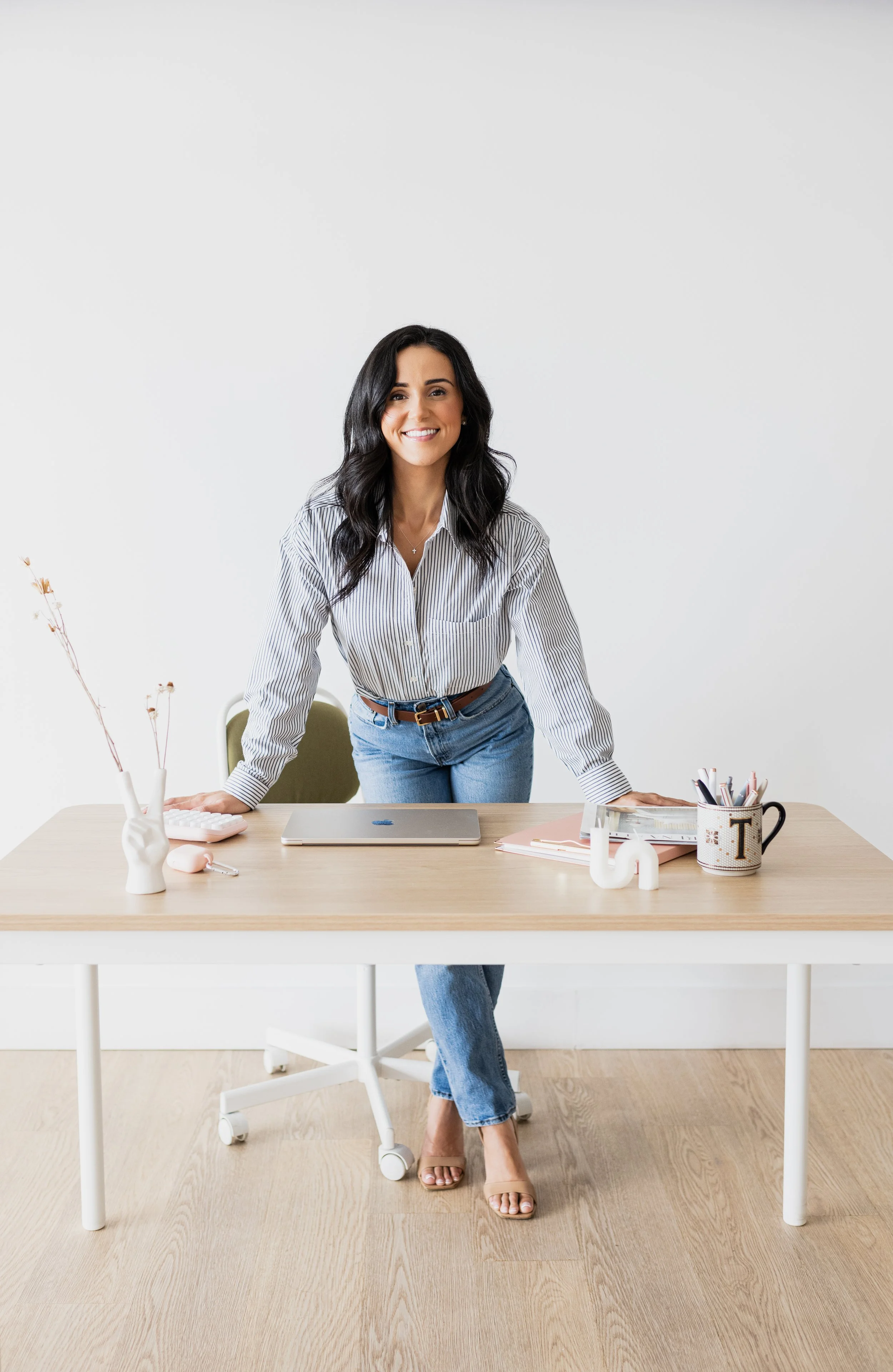 A woman with dark hair, wearing a striped shirt and jeans, leaning on a wooden desk with a laptop and office supplies, smiling at the camera.
