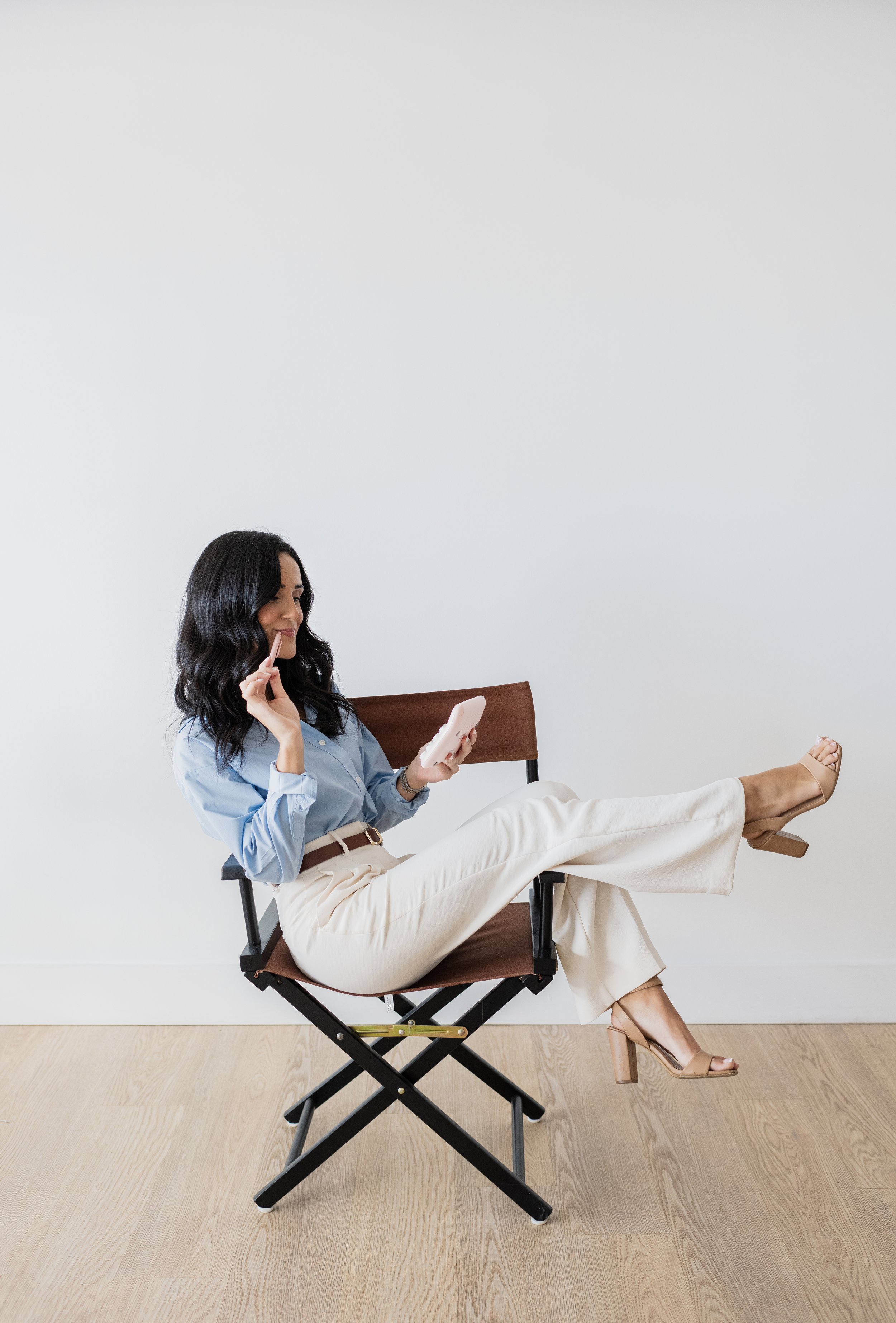 A woman with black wavy hair, wearing a light blue shirt and beige pants, sitting on a foldable chair with one leg resting on the other, looking at her smartphone and smiling in a minimalistic room with light wood flooring and plain white wall.
