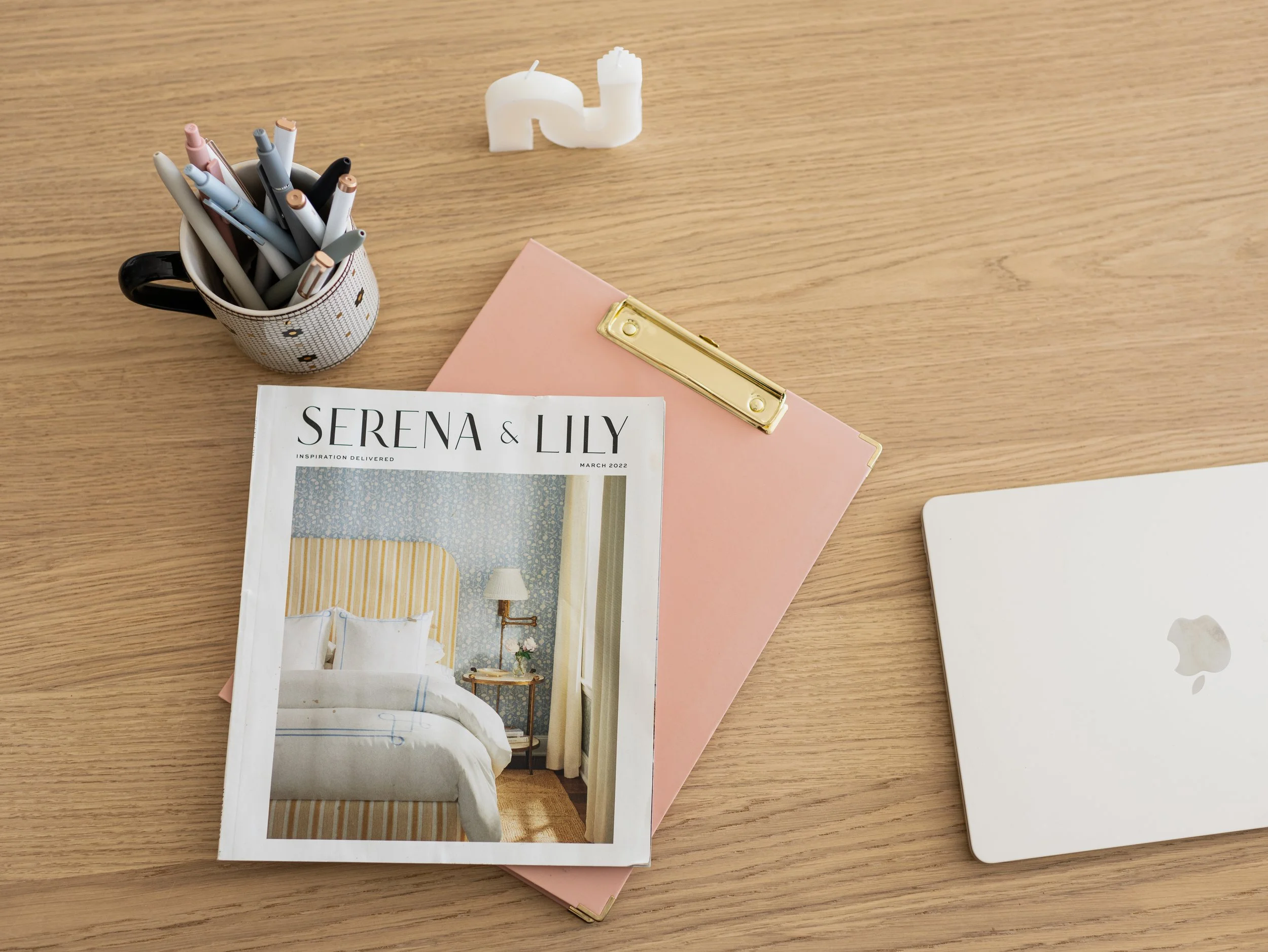 A wooden desk with a cup of black and pastel colored pens, a white candle, a pink clipboard with a gold clip, a pink notebook, a magazine titled "Serena & Lily" showing a bedroom, and a closed white Apple MacBook.