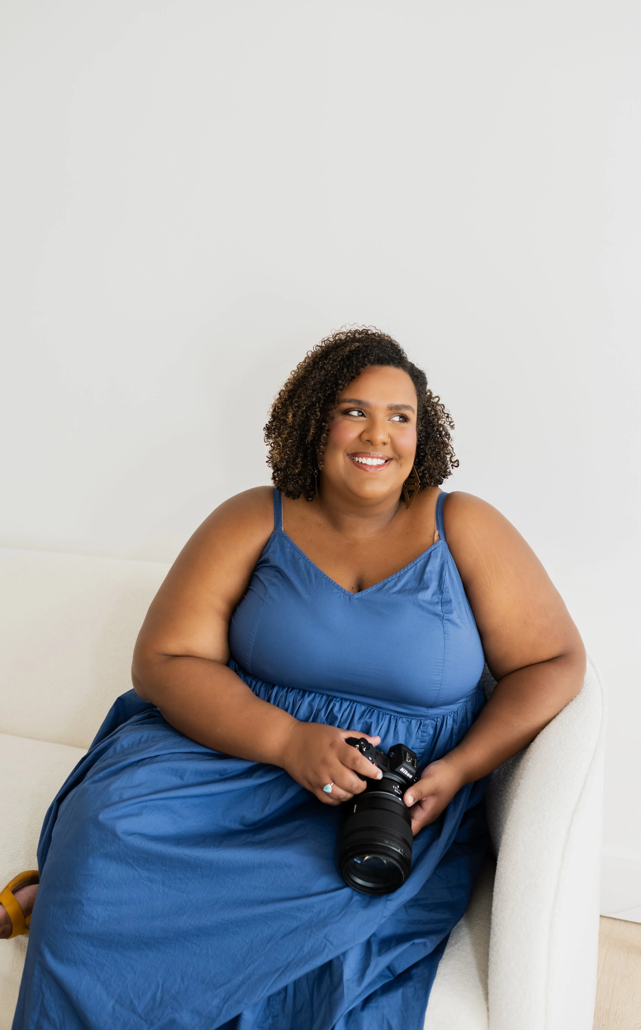A woman sitting on a white sofa, wearing a blue dress, holding a camera, smiling, with curly hair and earrings, against a plain white wall.