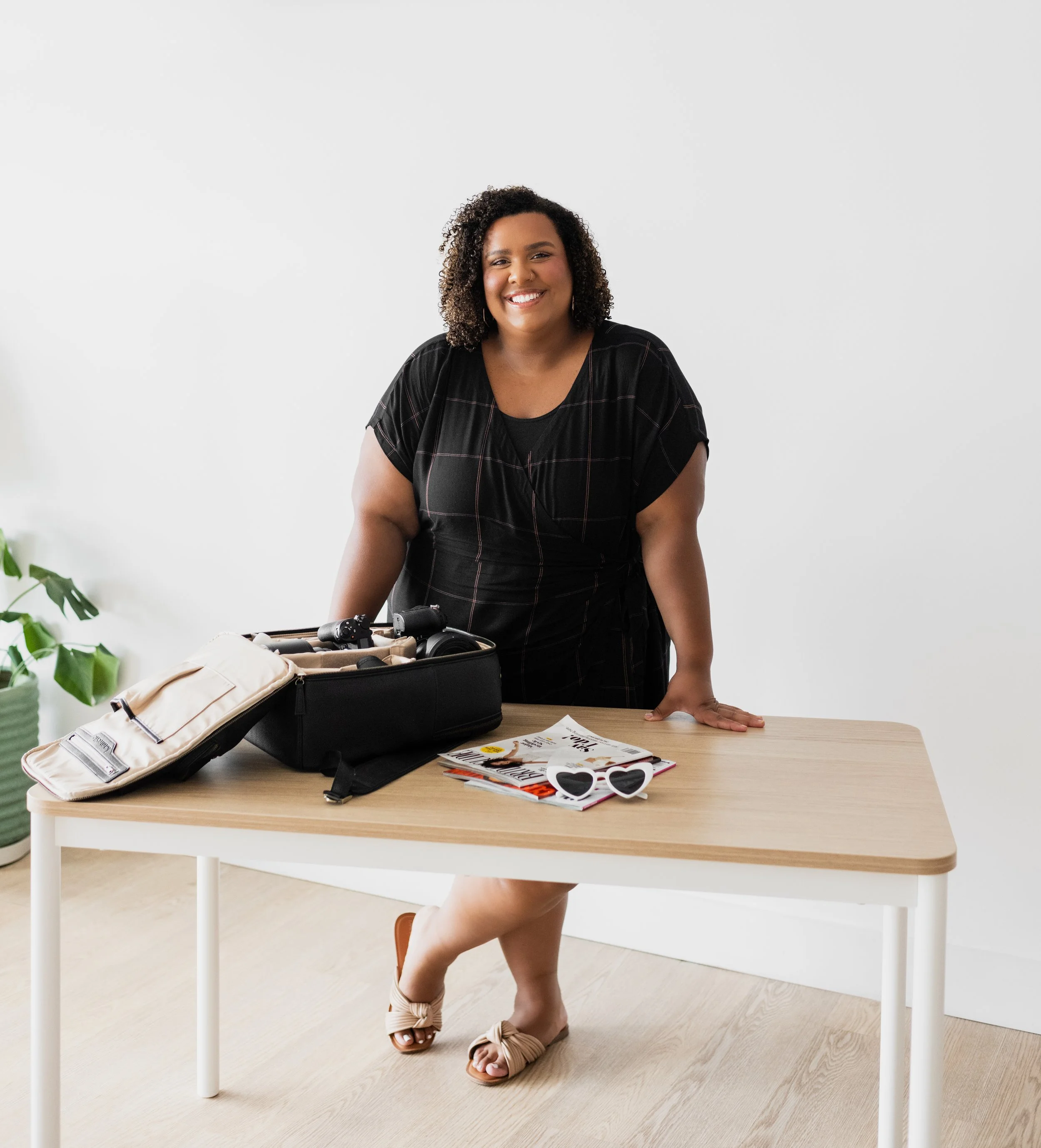 A smiling woman standing behind a wooden table with travel items, including magazines, a pair of heart-shaped sunglasses, a backpack, and a bike helmet, in a bright room with a white wall and a green plant.