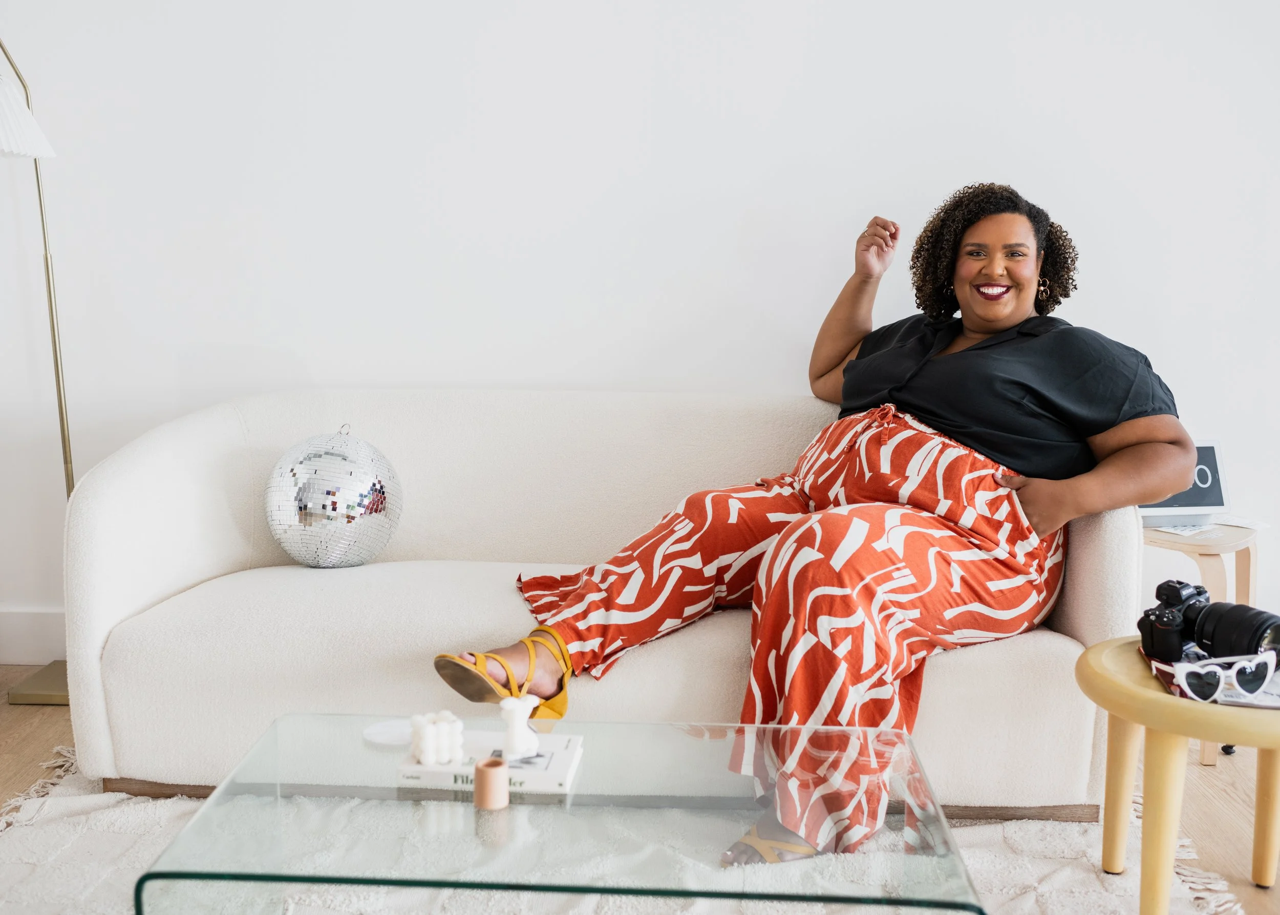 A woman with curly hair smiling and sitting confidently on a white sofa in a modern, minimalistic living room with a disco ball, camera, sunglasses, and a small wooden side table.