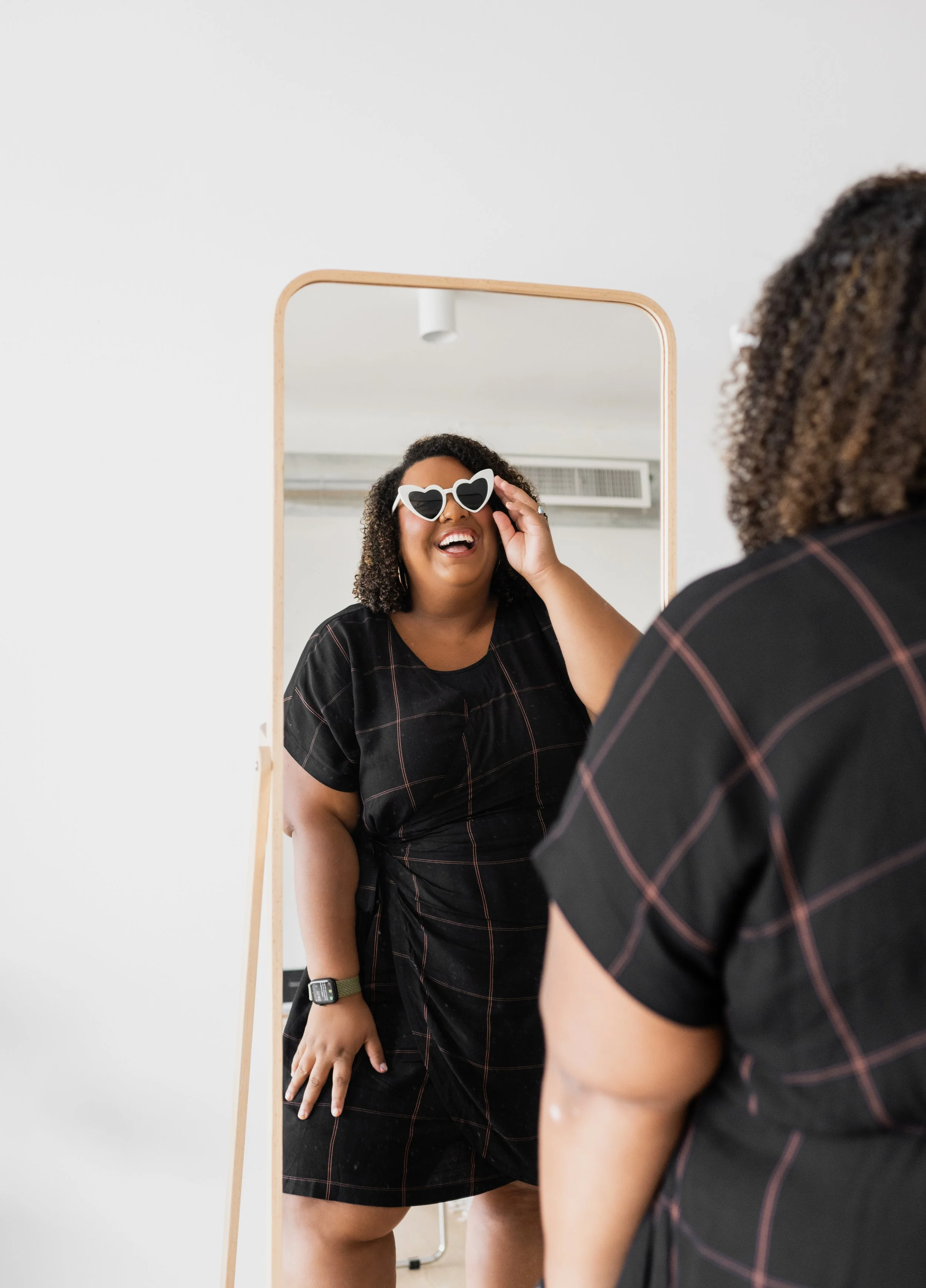 A woman with curly hair trying on heart-shaped sunglasses, smiling and looking at herself in a mirror.