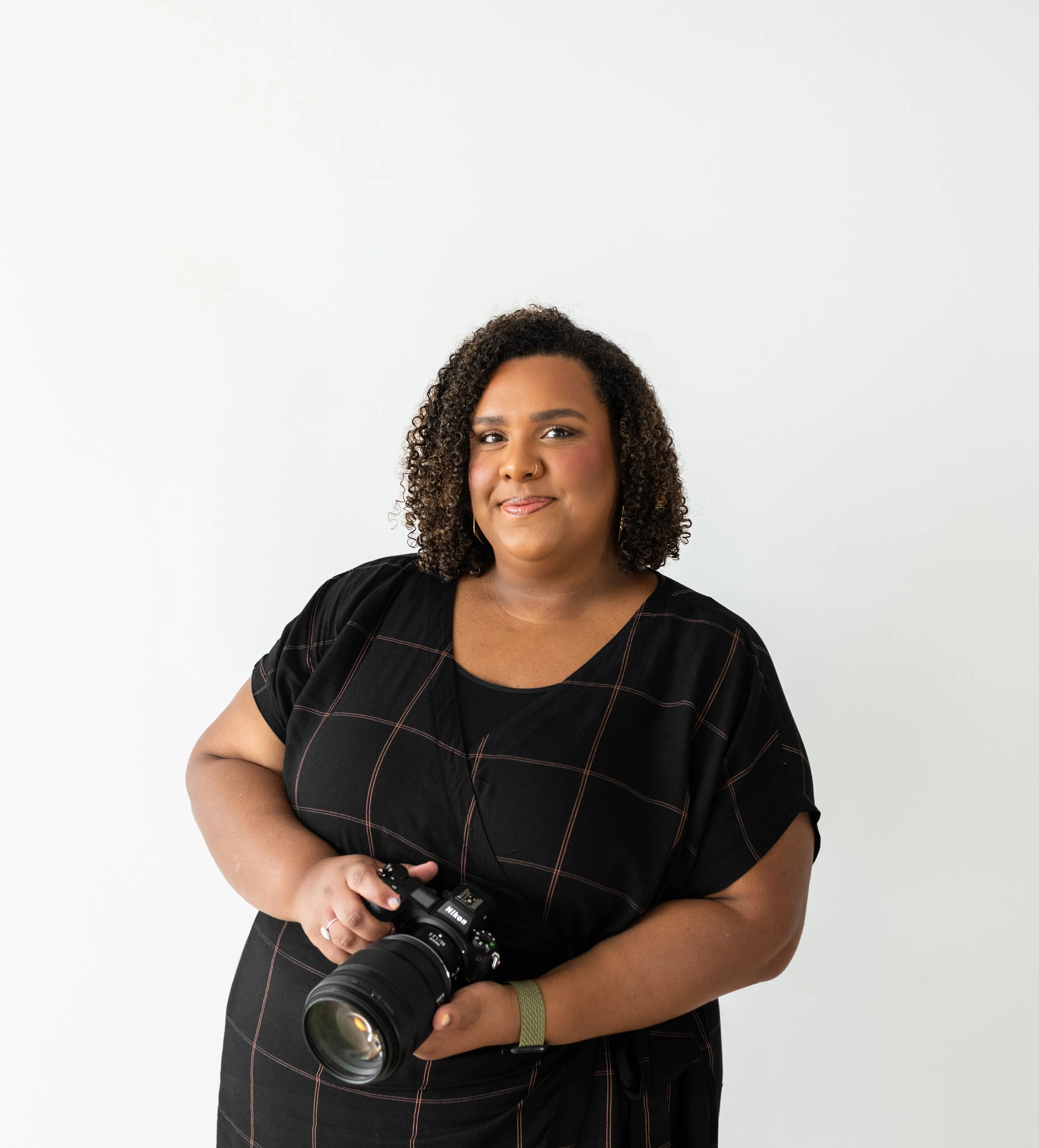 Woman with curly hair holding a camera, posing against a plain white background.