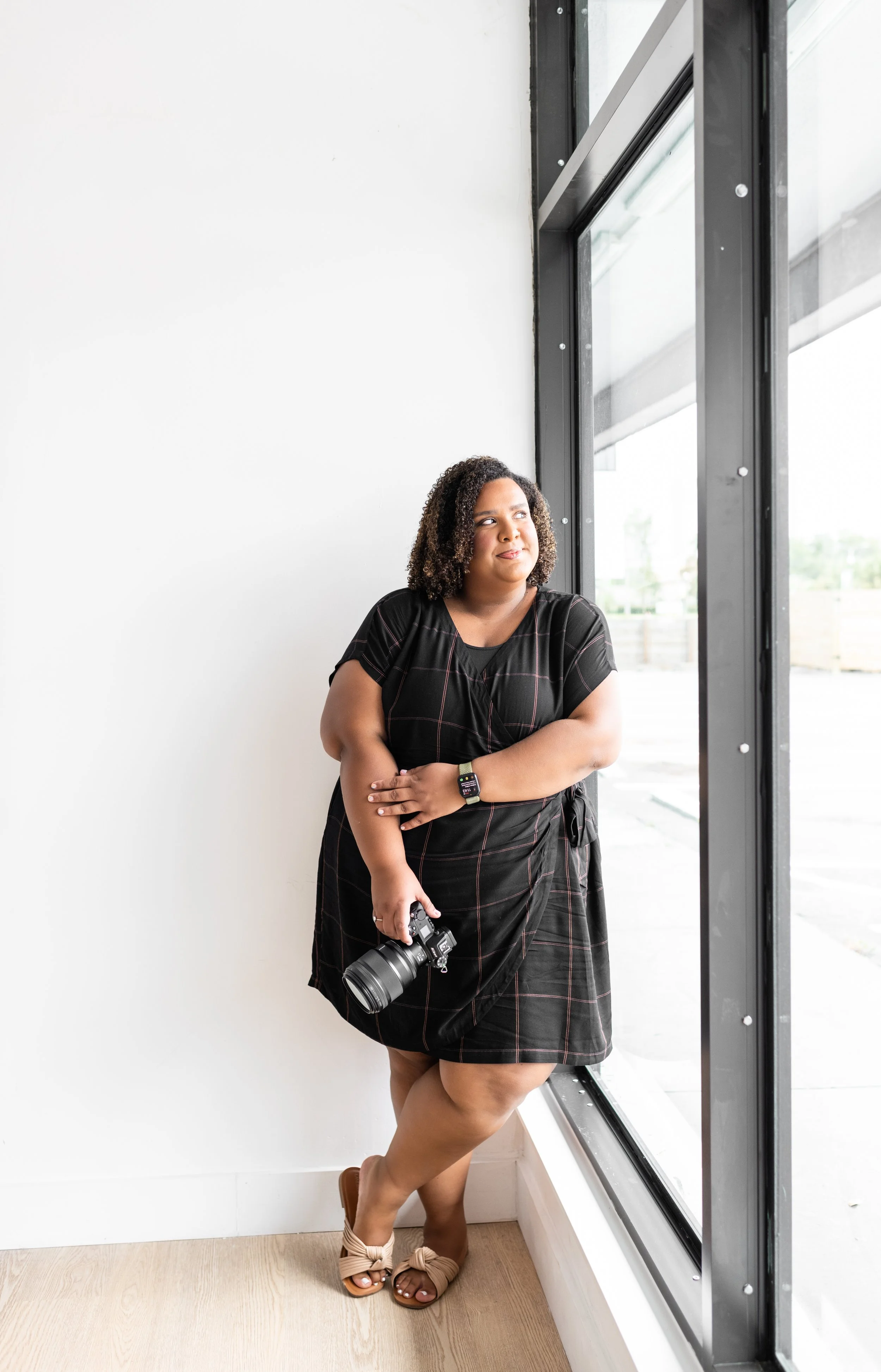 A woman standing by a large window, holding a camera in one hand, wearing a black dress, a smartwatch, and sandals with bows, looking thoughtfully outside.