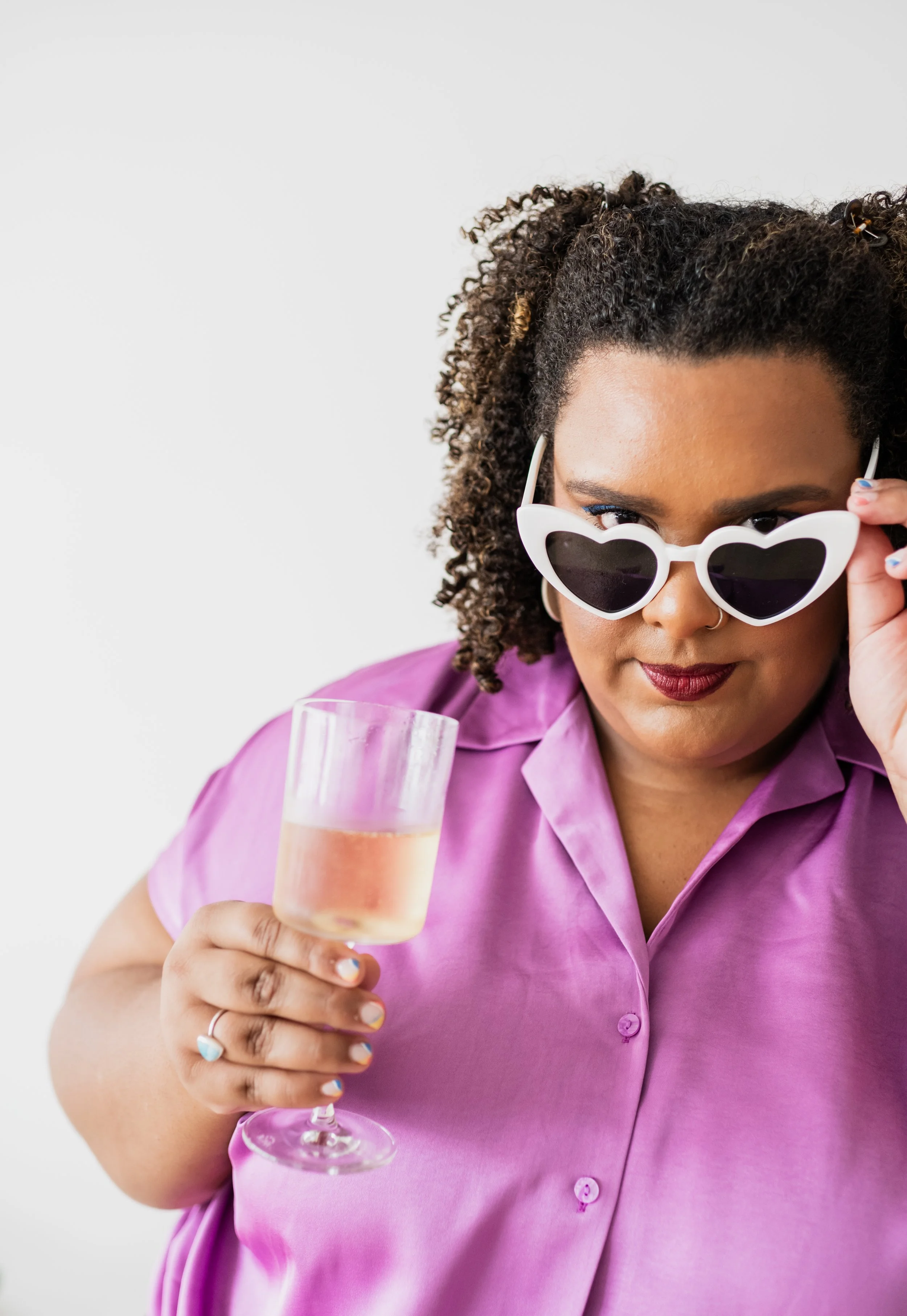 A woman wearing heart-shaped sunglasses and a purple shirt, holding a glass of champagne, looking at the camera.