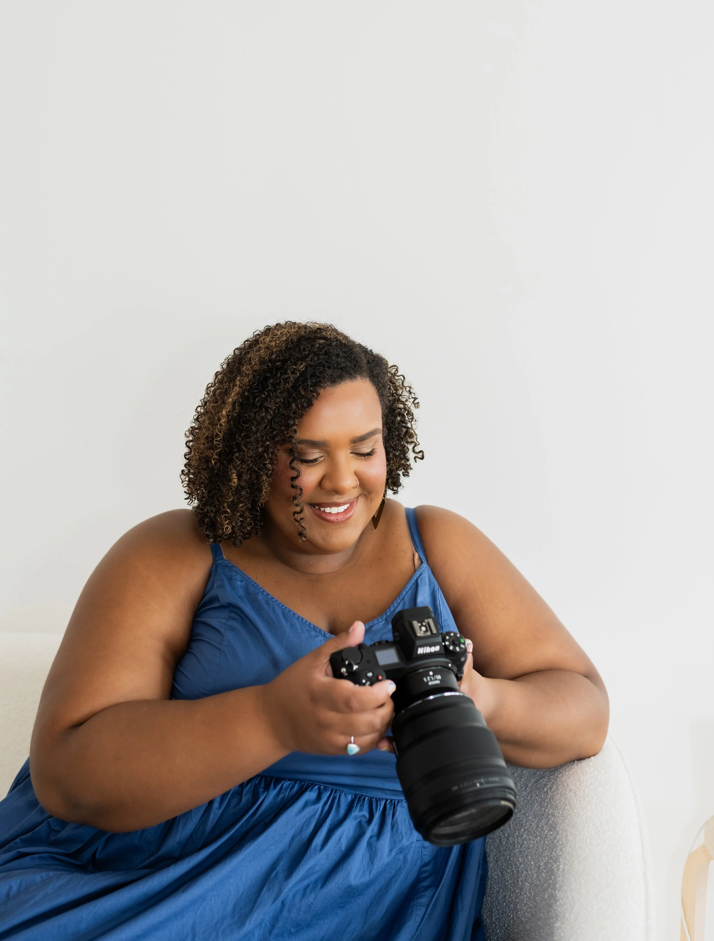 A woman with curly hair wearing a blue dress sitting on a light-colored sofa, smiling and looking at a professional camera in her hands, against a plain white wall.