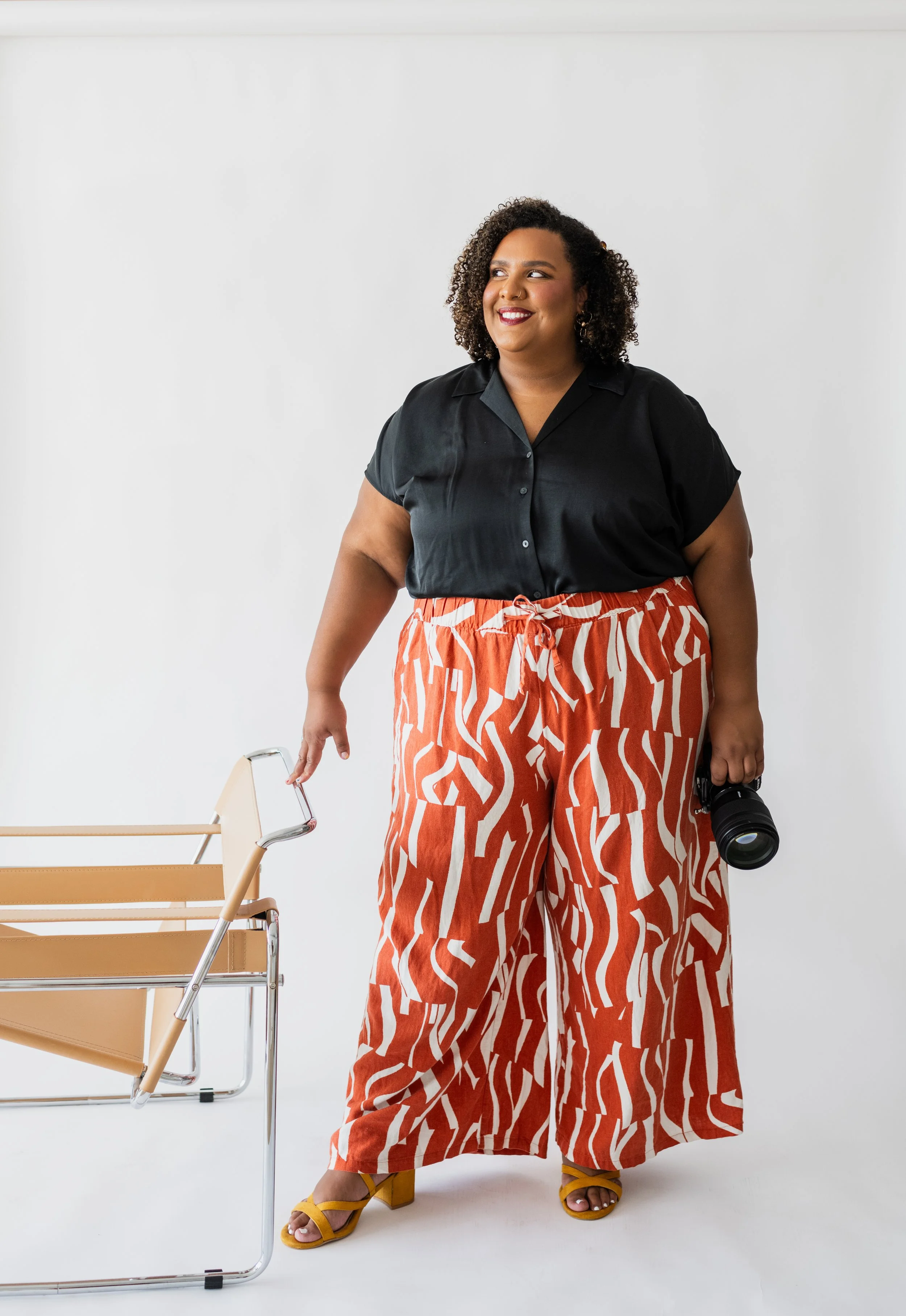 A woman with curly hair wearing a black shirt and orange patterned wide-leg pants, holding a camera in her right hand and standing next to a tan lounge chair against a white background.