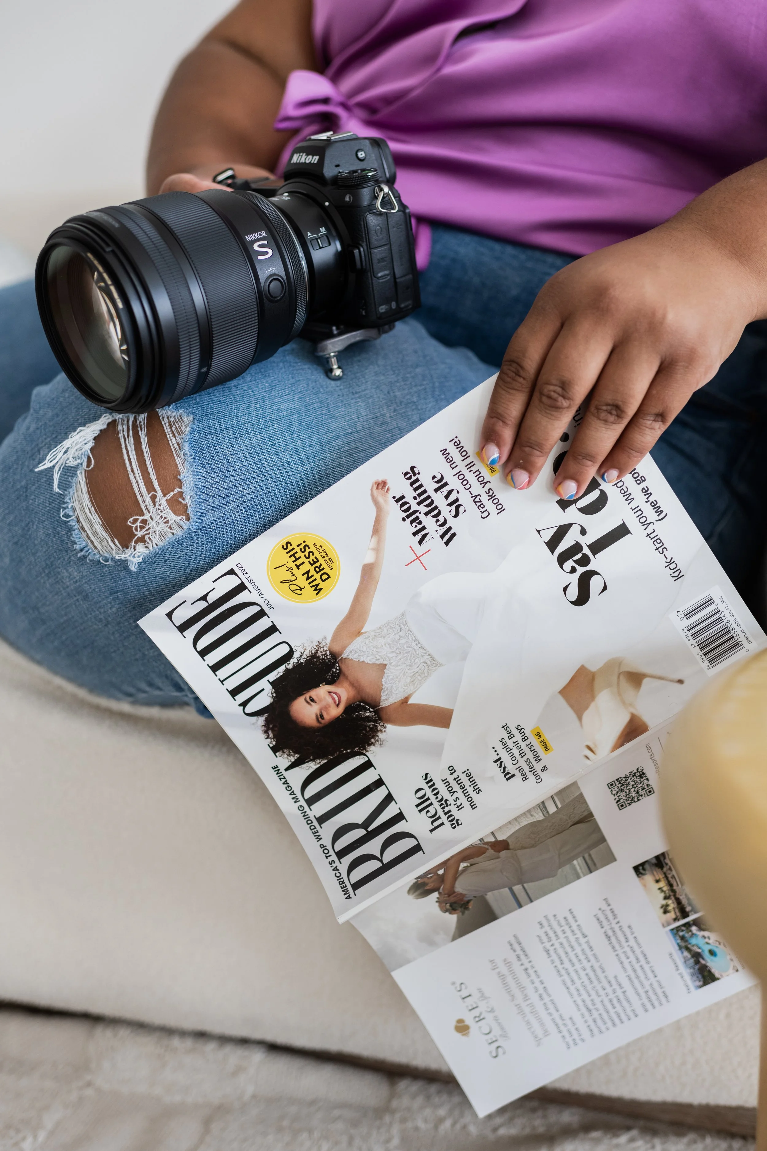 A person sitting on a beige sofa holding a magazine with a Nikon camera resting on their knee. The person is wearing distressed jeans and a purple top with painted nails.