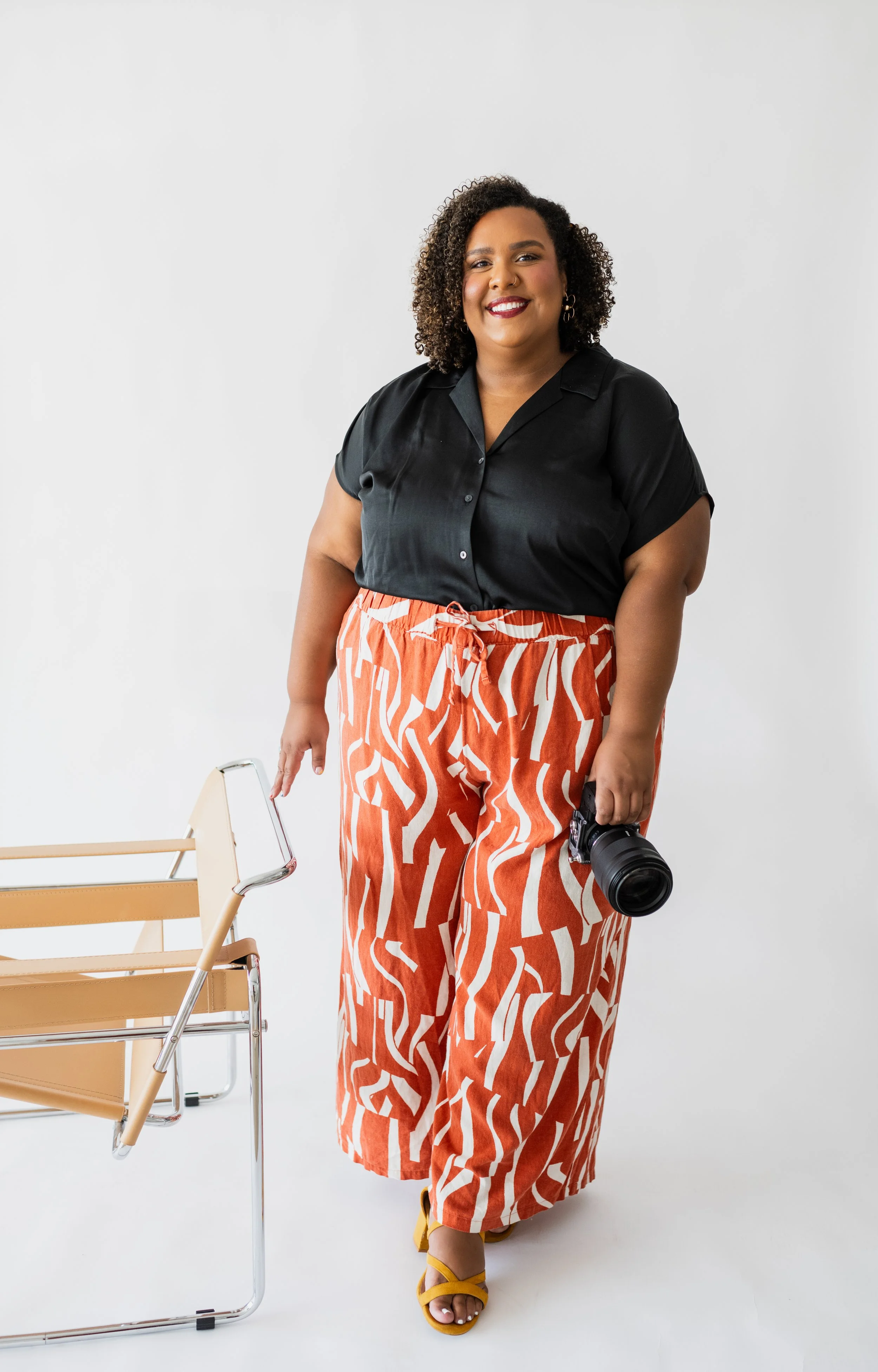 A woman with curly hair, wearing a black shirt and orange patterned pants, standing against a white background, holding a camera in her right hand, and a chair beside her.